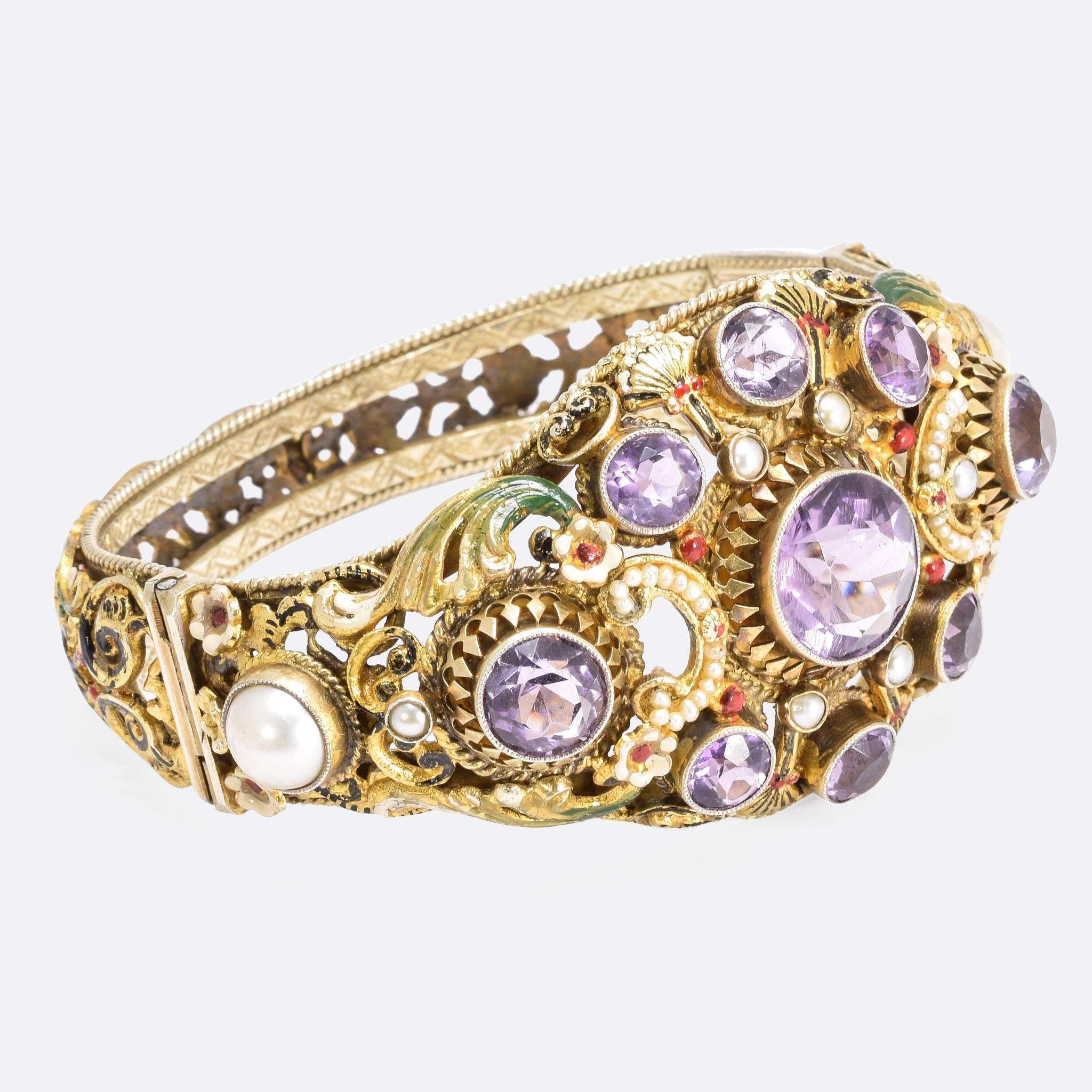 A stunning ornate enamelled bangle modelled in silver gilt. With intricate floral and foliate openwork throughout, the front is set with 9 well matched coloured amethysts, and pearls surrounding the principal stone. The colour pallet and style are