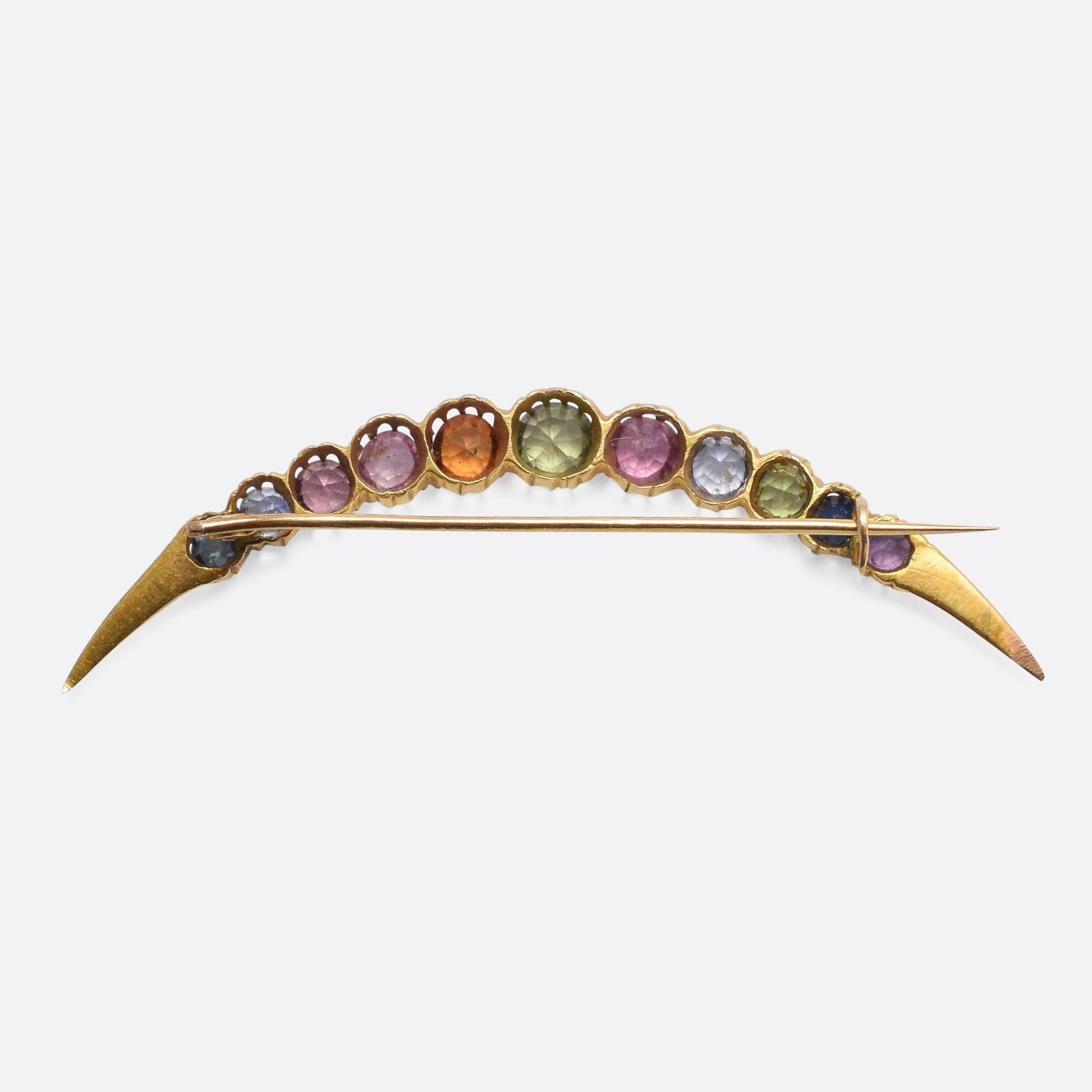 A gorgeous Victorian harlequin crescent brooch, set with a rainbow of coloured gemstones in scalloped claw settings. The stones include a central demantoid garnet, flanked by: blue and pink topaz, hessanite garnet, blue and pink sapphires, and