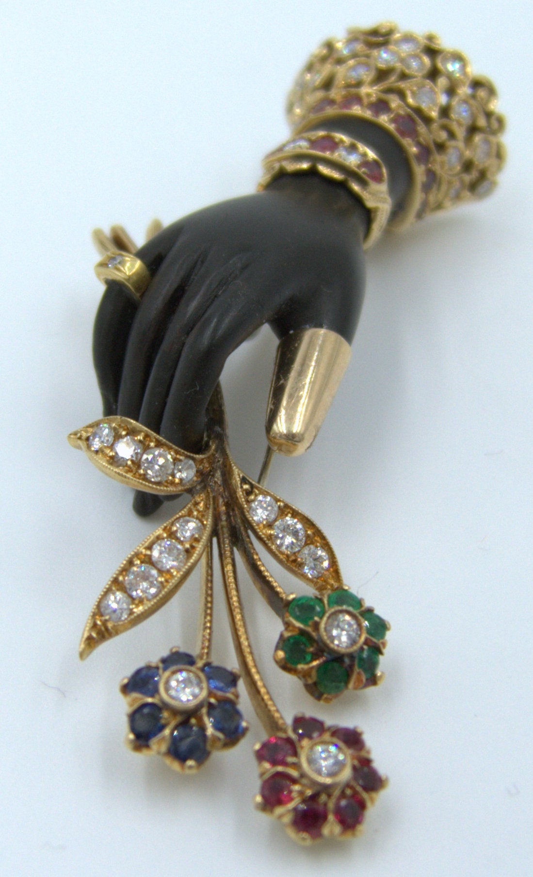 A true departure from the typical Nardi moretti brooch, this delightful matte black agate hand adorned with a gold, ruby and diamond set lace cuff holds a sapphire, emerald, ruby, and diamond floral spray.   A jeweled ring adorns one finger, and a