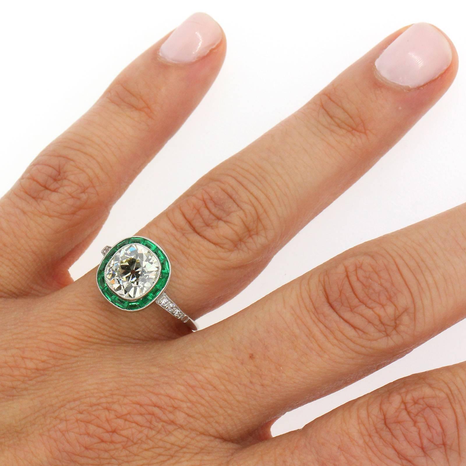 A refined recreation of a style gone by, this beautiful ring features a two and half carats Old Cushion Cut Diamond of warm color and high clarity.  The stunning diamond is surrounded by calibrated deep green Emeralds, and six Old European Cut