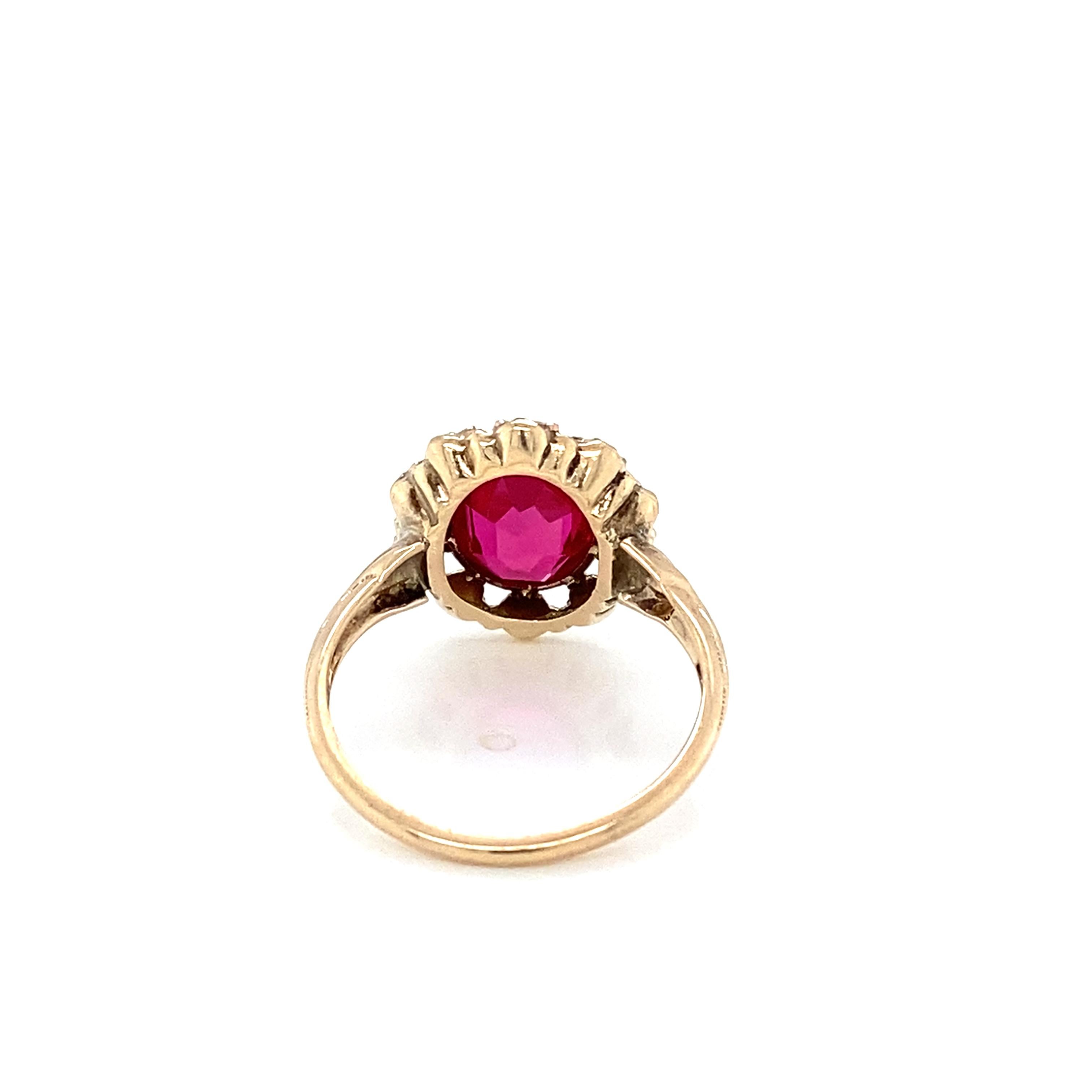 Dieser bezaubernde Art-Déco-Ring aus 10 Karat Gelbgold mit einem Hauch von Roségold enthält einen funkelnden, facettierten, 10 mm x 8 mm großen, ovalen, brillanten, roten, synthetischen Rubin, der in einer Lünette gefasst und von einem Kranz aus