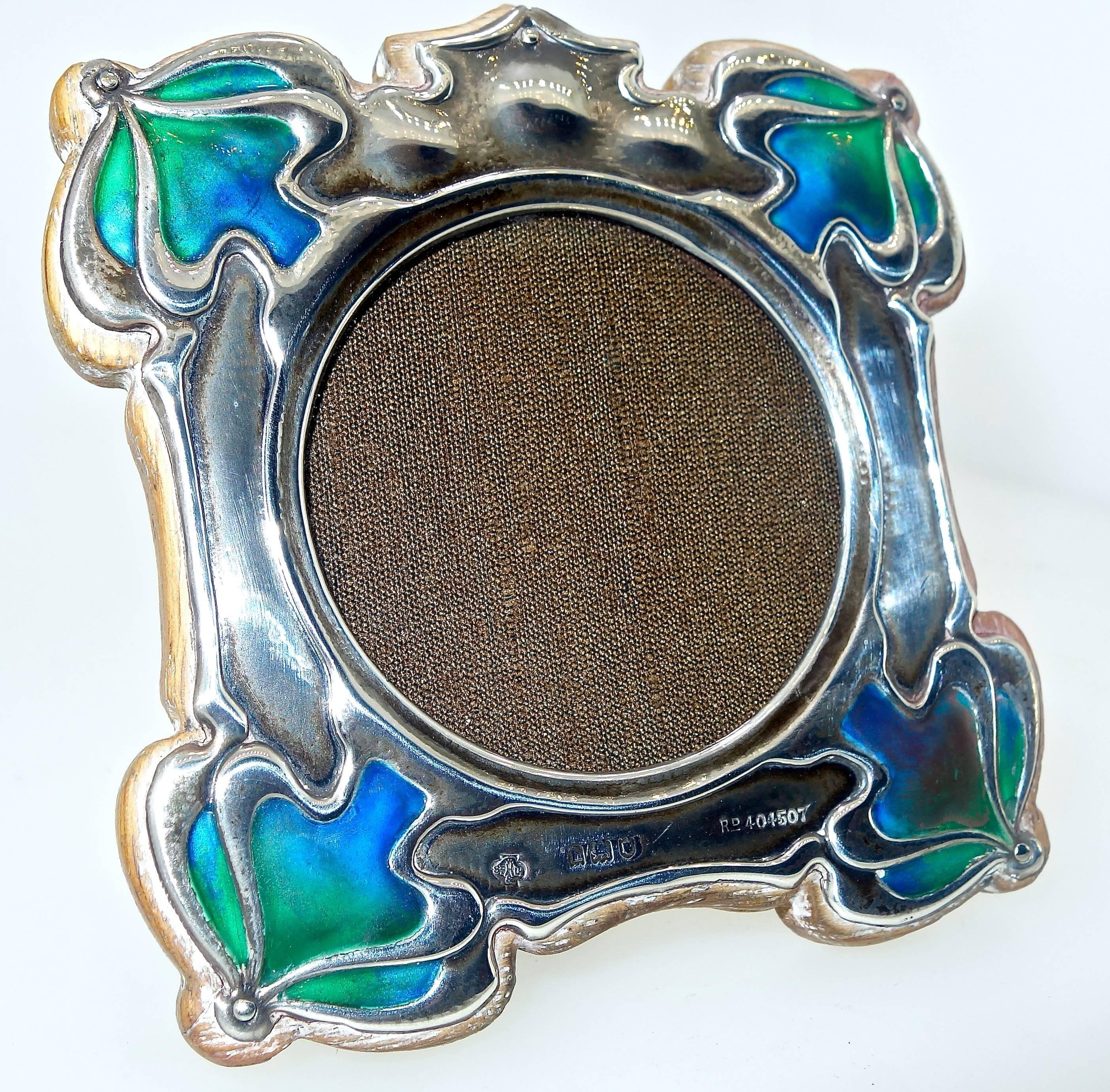 Shaded peacock color enamel accent the corners of this antique sterling silver frame.  This period piece bears the English marks of a small h, a lion and a shield.  The piece bears the maker's mark and is numbered.  The  back and easels are the