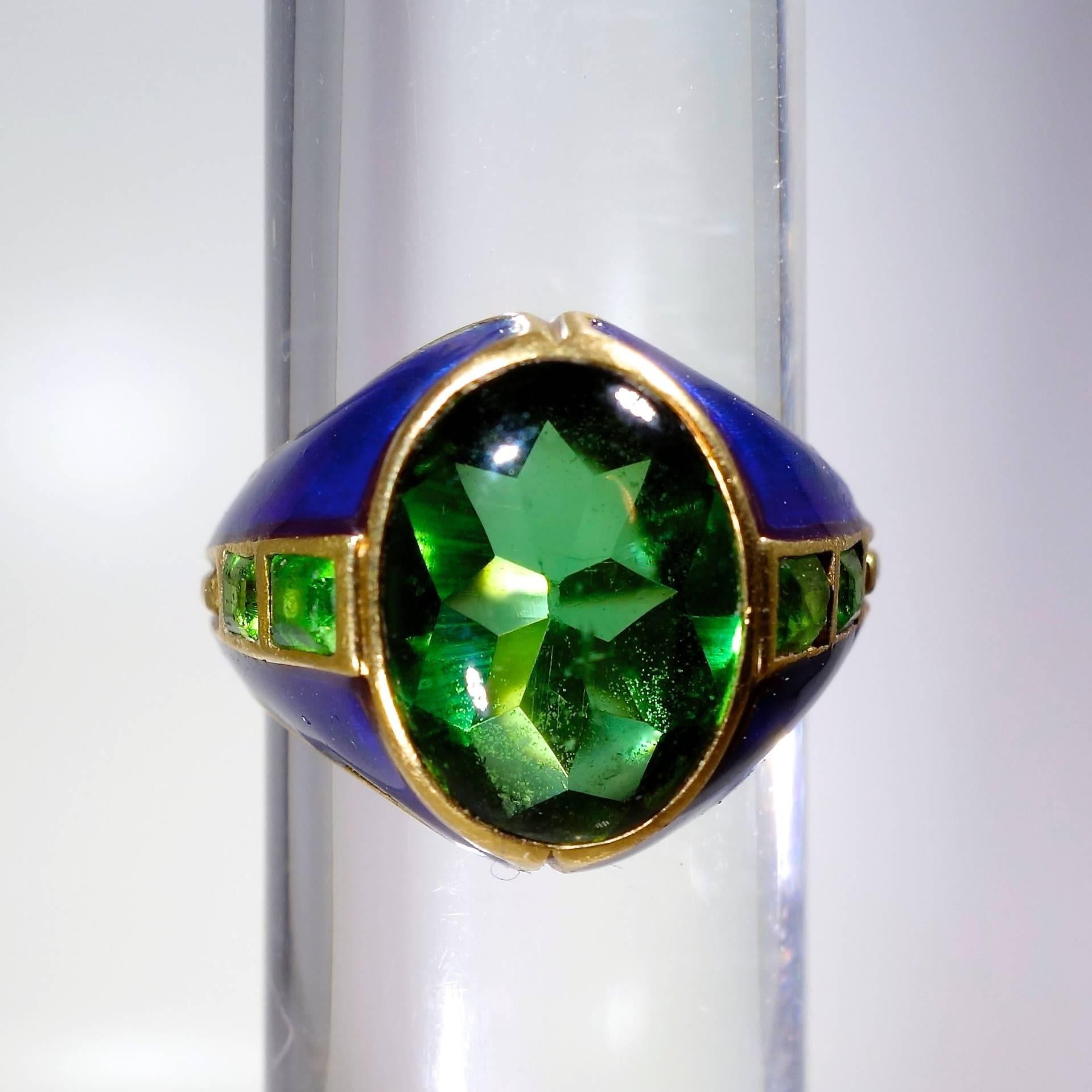 The  center stone is a buff top faceted bottom Peridot, 11.6 by 8.8 mm.,  bright cobalt blue iridescent enamel, 4 square cut Demontoid garnets, and a gold bead design which further accent the sides.  The book, Louis Comfort Tiffany by John Loring,