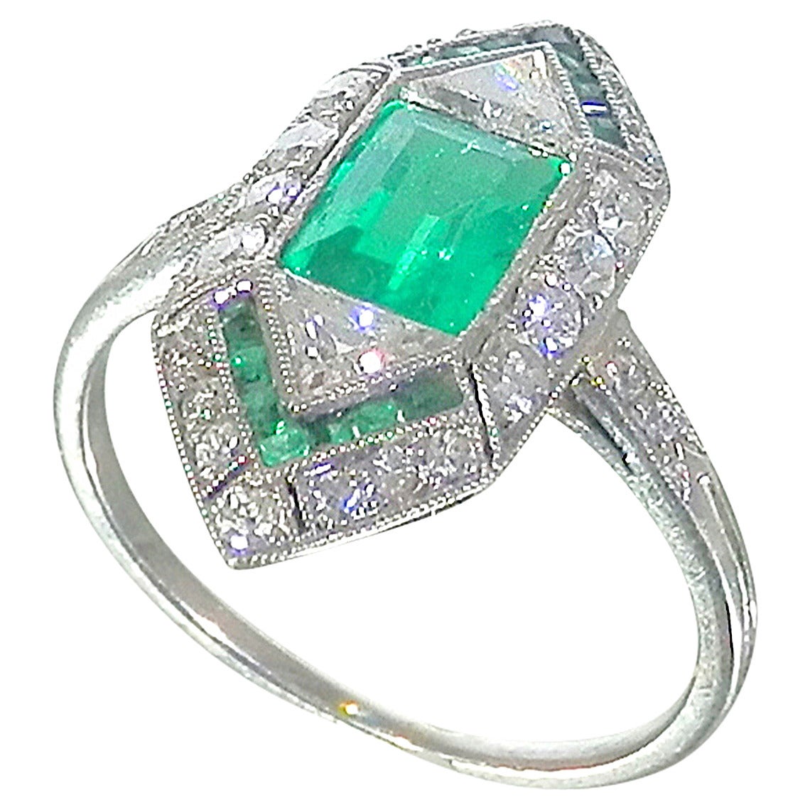 In excess of 1 ct., the emerald in this ring is Colombian displaying a fine medium to light bright green color.  There are 14 caliber cut smaller matching emeralds.  There are 2 old triangle cut diamonds on either side of the center emerald.  There