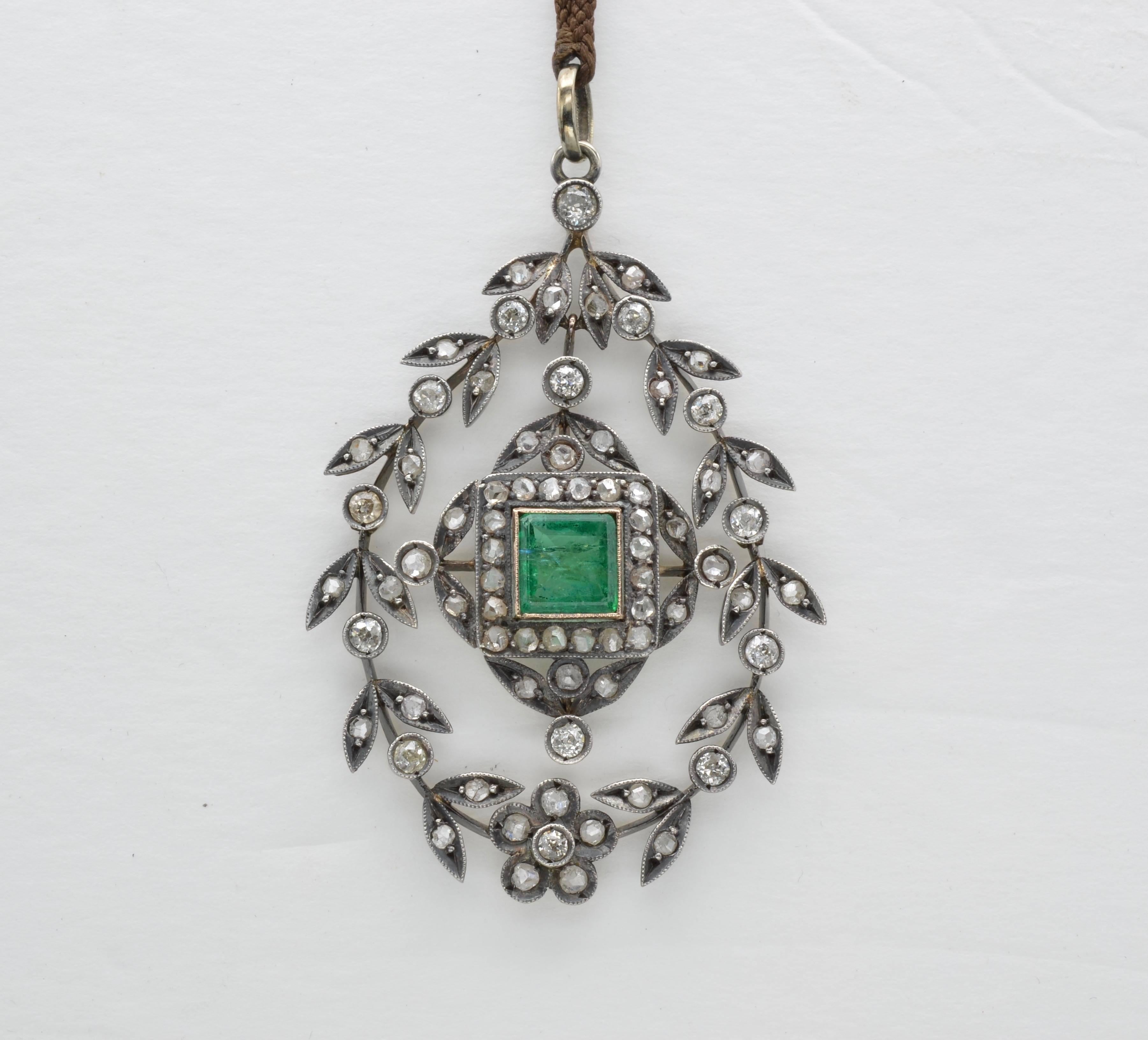 This large wreath-like pendant is full of old mine cut diamonds and a hinge in the middle to create a swinging movement.  A large bright green emerald glows in the center with horizontal inclusions and historical allure. It is on a braided cord so