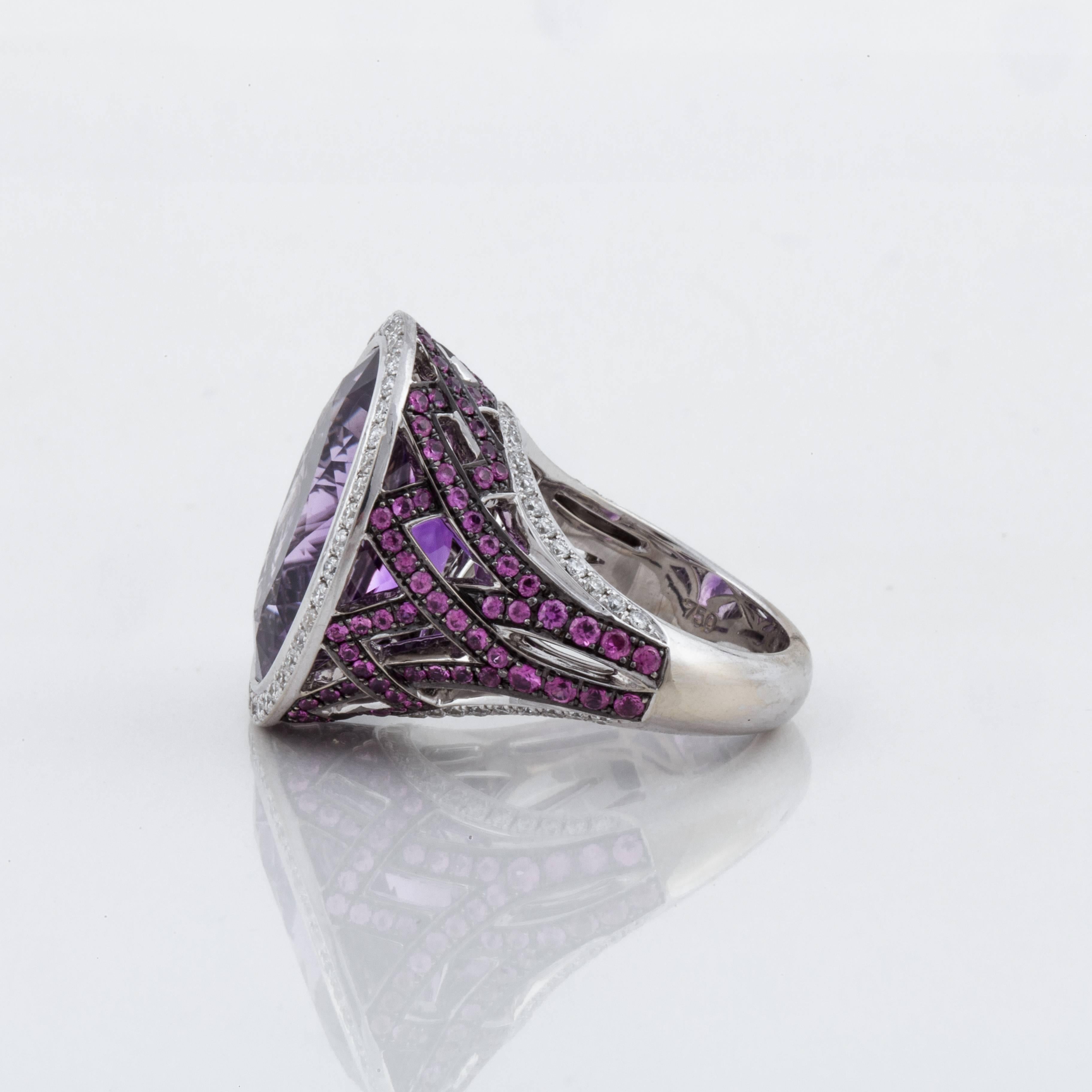 Asprey ring in 18K white gold featuring an oval amethyst framed by round diamonds.  The amethyst is 12 carats with 0.90 carats in diamonds.  There are also pink sapphires along the sides of the ring that total 1.95 carats.  Marked 