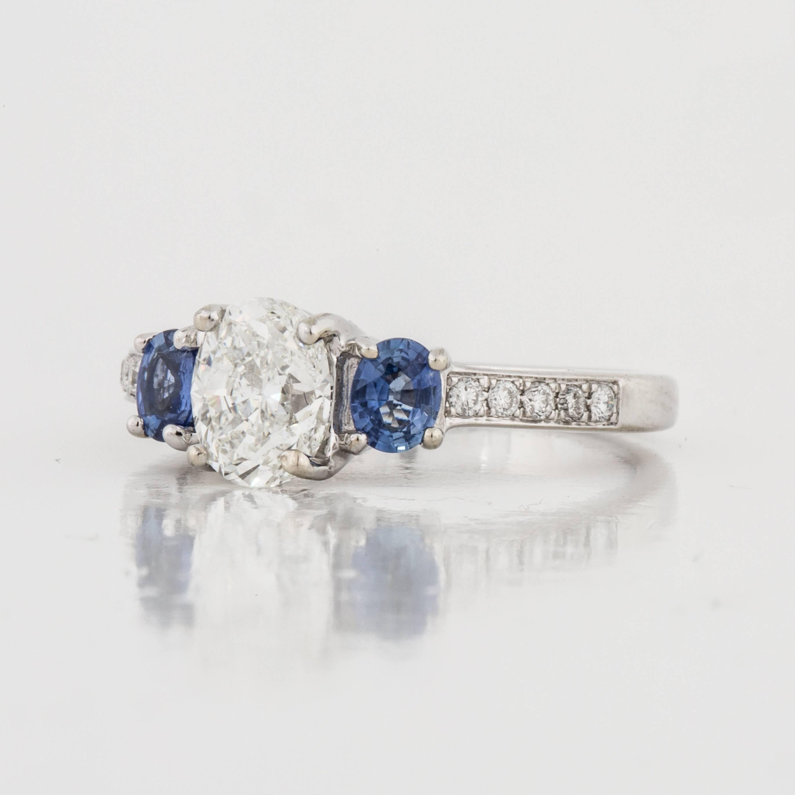 Three-stone diamond and sapphire engagement ring composed of 14K white gold.  The center oval diamond is 0.94 carats, H-I color and SI1-SI2 clarity.  On each side of the central diamond are two oval shaped blue sapphires that total 0.40 carats. 