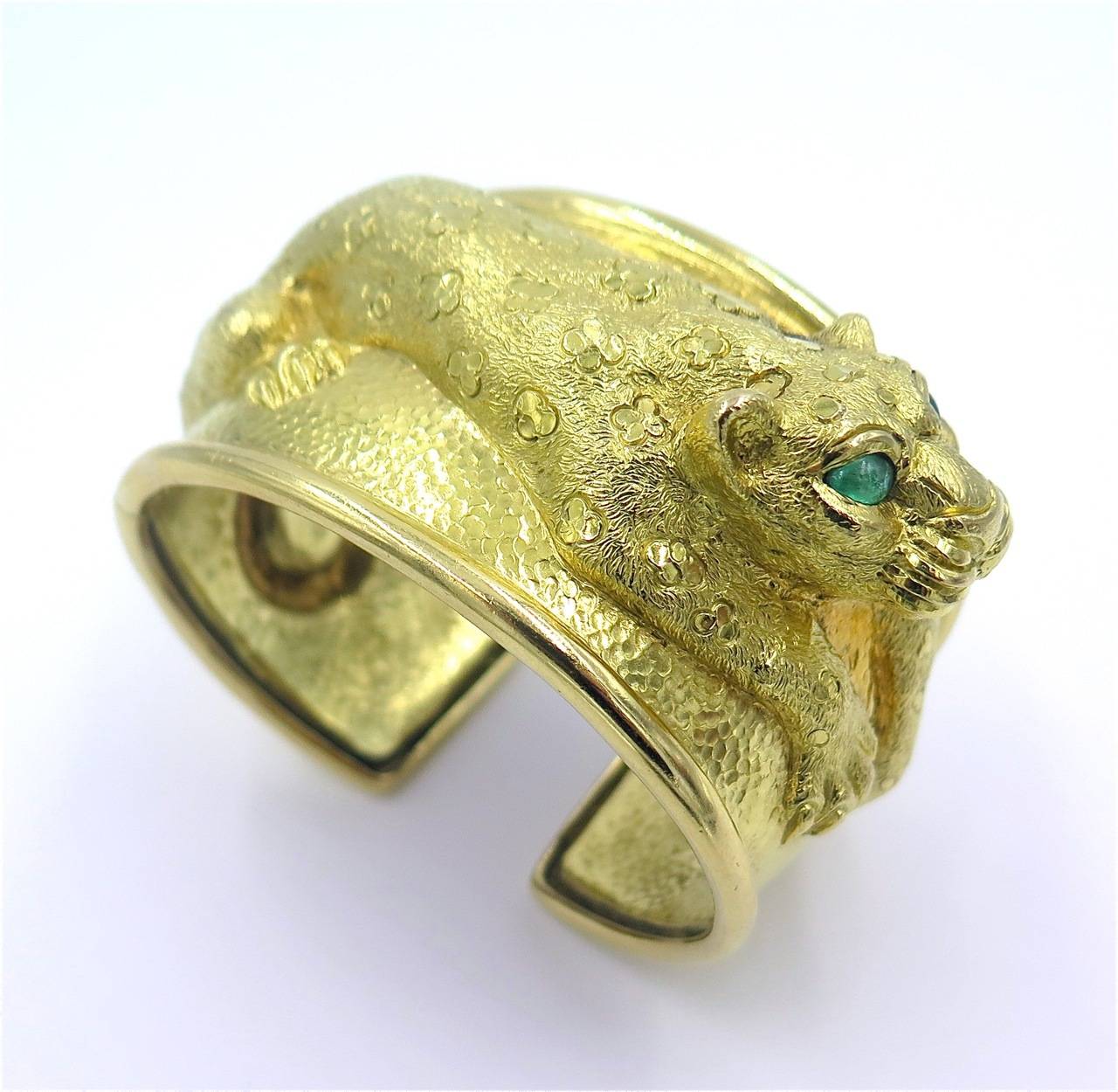 An 18 karat yellow gold and emerald “Leopard” cuff bracelet, David Webb, Circa 1980.  Signed WEBB 18K.  The wide textured gold cuff depicts a crouching leopard, the eyes set with pear-shaped emeralds weighing approximately .75 carat total.  The