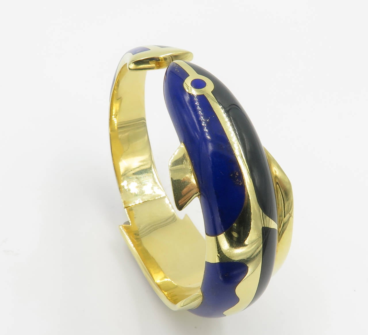An 18 karat yellow gold, lapis lazuli and black jade hinged bangle bracelet, Tiffany & Co., Circa 1975.  The bracelet is designed in the form of a fish with gold fins and tail, inlaid with sections of black jade and lapis lazuli.  The bracelet has a