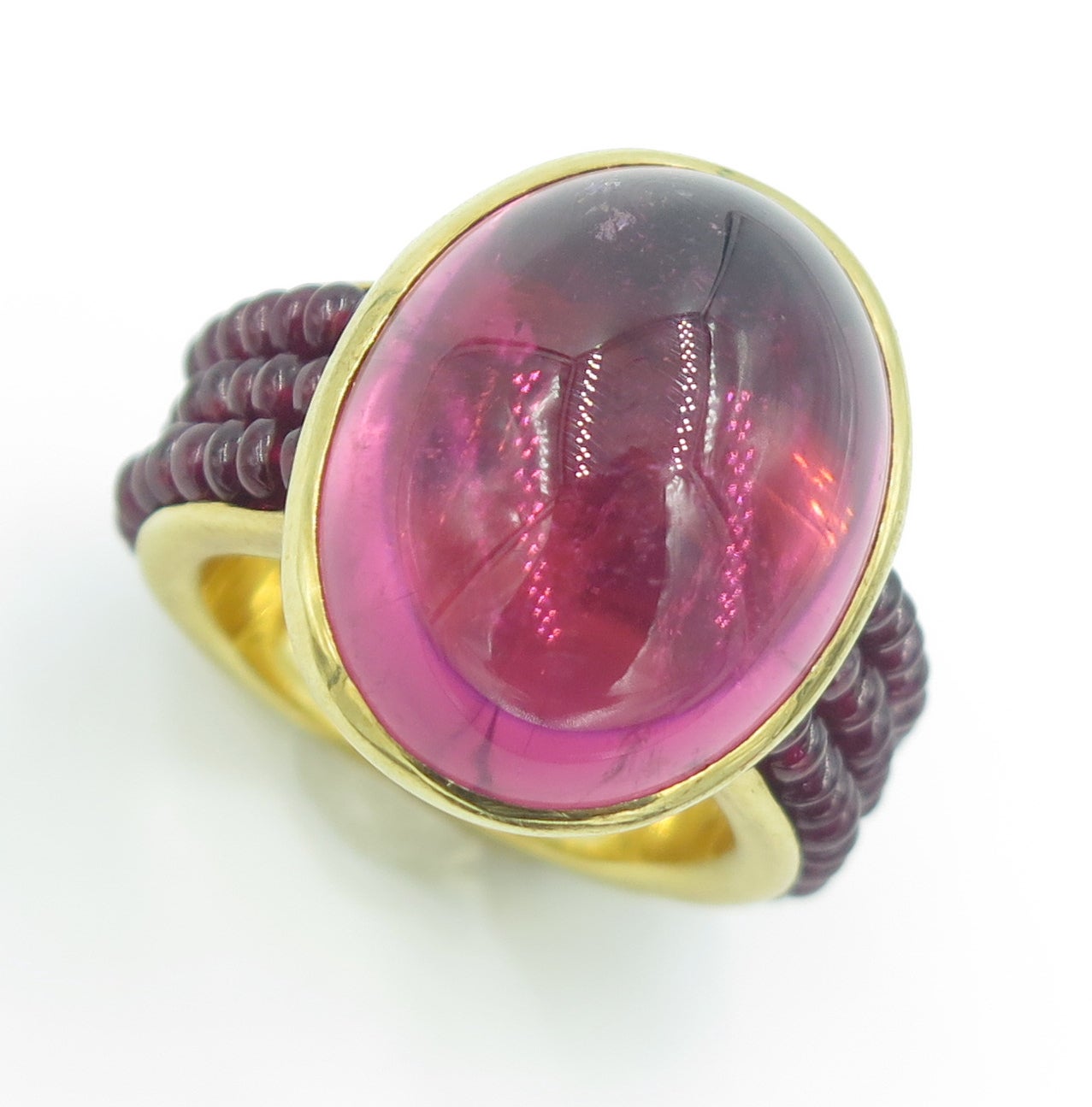 An 18 karat yellow gold, pink tourmaline and ruby bead ring, McTeigue & McClelland.  The ring is collet set with an oval cabochon pink tourmaline weighing approximately 21.6 carats.  The shoulders are each set with 3 strands of ruby beads.  The ring
