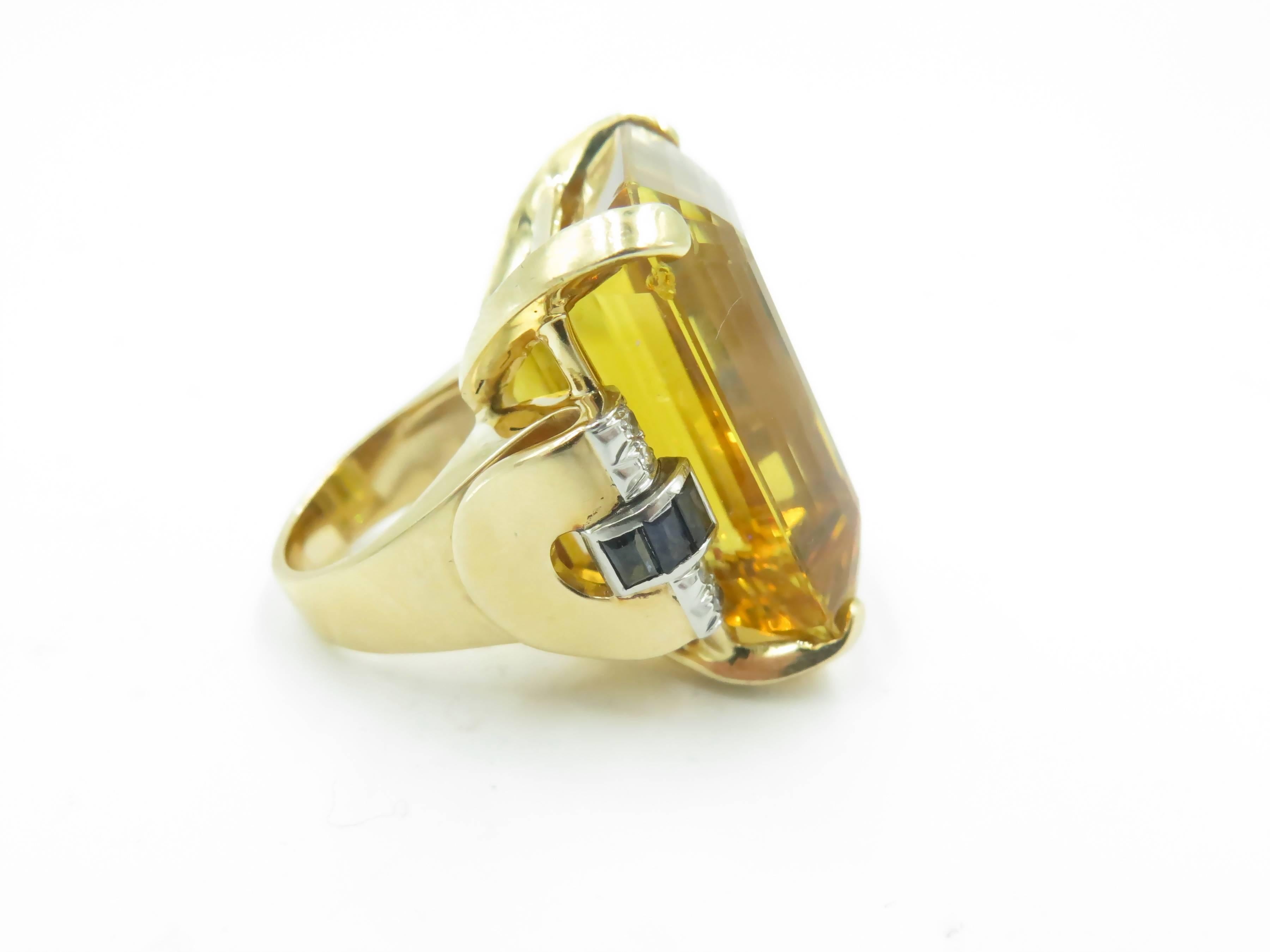 A 14 karat yellow gold citrine, sapphire and diamond ring. Circa 1940. Set with an emerald cut citrine, measuring approximately 29.00 x 21.00 x 16.00mm, and weighing approximately 69.00 carats. flanked by calibre cut sapphire and circular cut