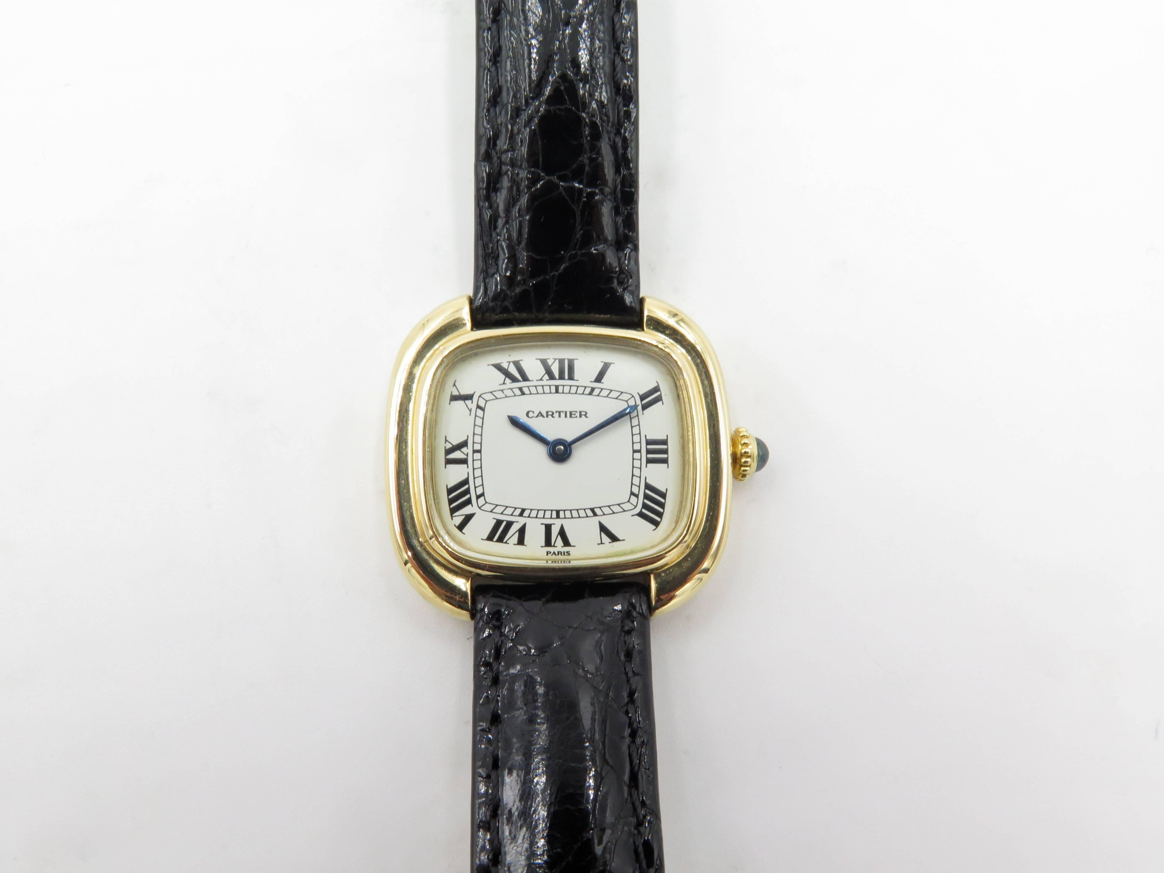 An 18 karat yellow gold ladies watch, Cartier. Paris.  Known as the Ellipse model. Circa 1975. Of mechanical movement, the white dial of square outline, with Roman numerals and blued steel hands and cabochon sapphire crown, joined by a black leather