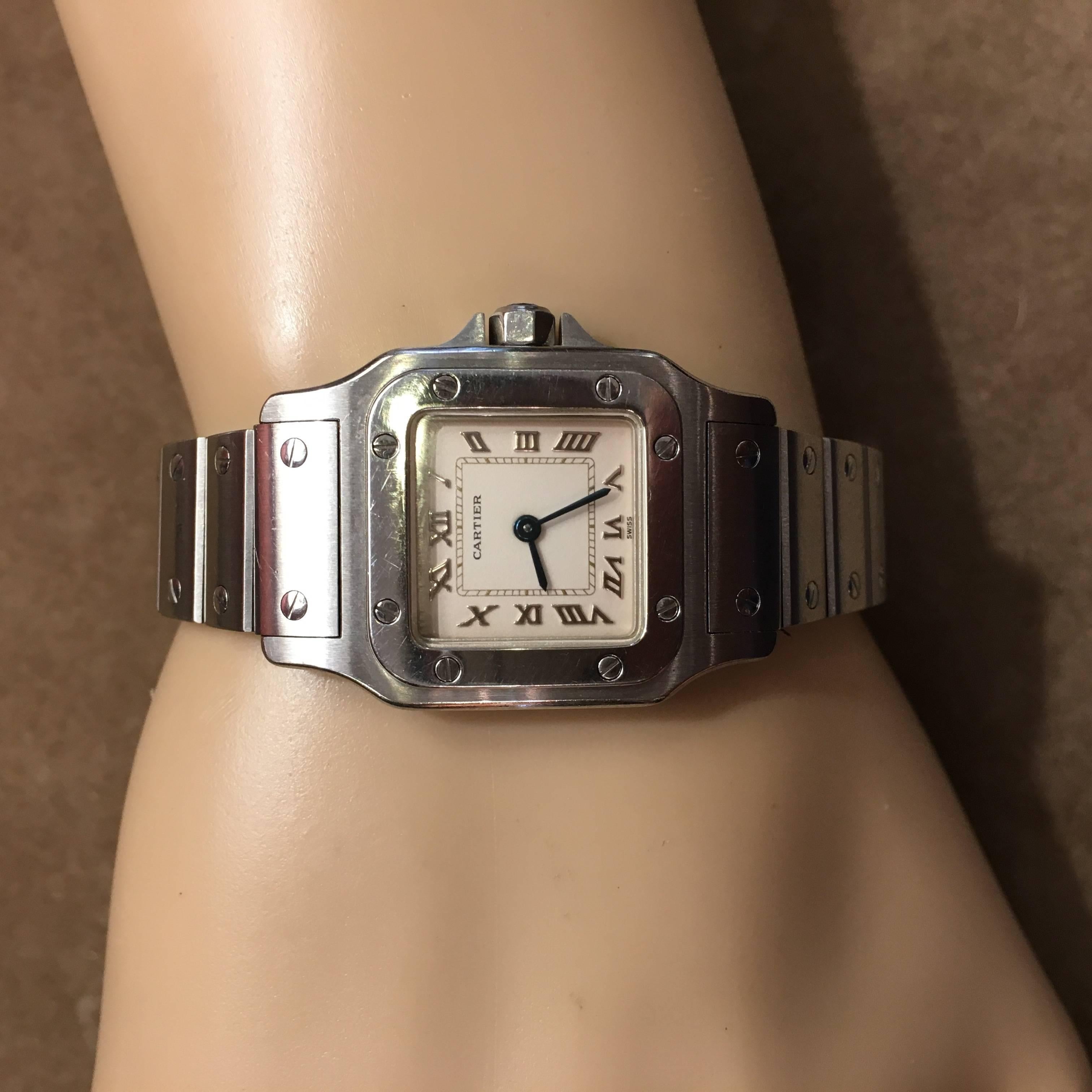A stainless steel ladies Santos watch. Cartier. Of movement, the square white dial with Roman numerals and blued steel hands. Joined by a matte steel bracelet with deployant buckle, bezel and bracelet enhanced by screws. Numbered 9057930 13876.