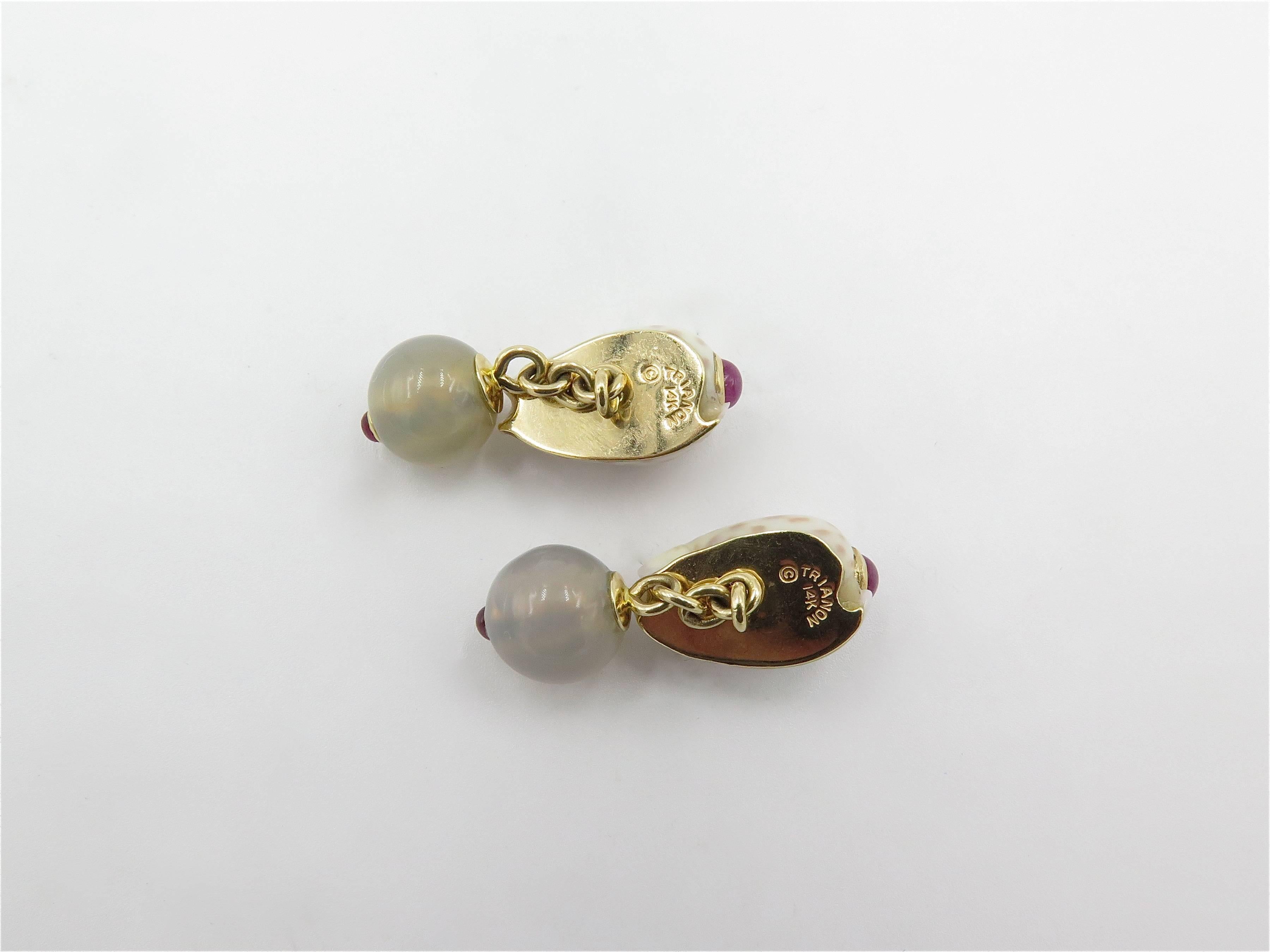 A pair of 14 karat shell and gem stone cufflinks. Trianon. Circa 2000. Each set with a mottled cream seashell studded with round cabochon rubies, joined by a chain to a round bluish gray chalcedony bead, measuring approximately 10.00mm, enhanced by
