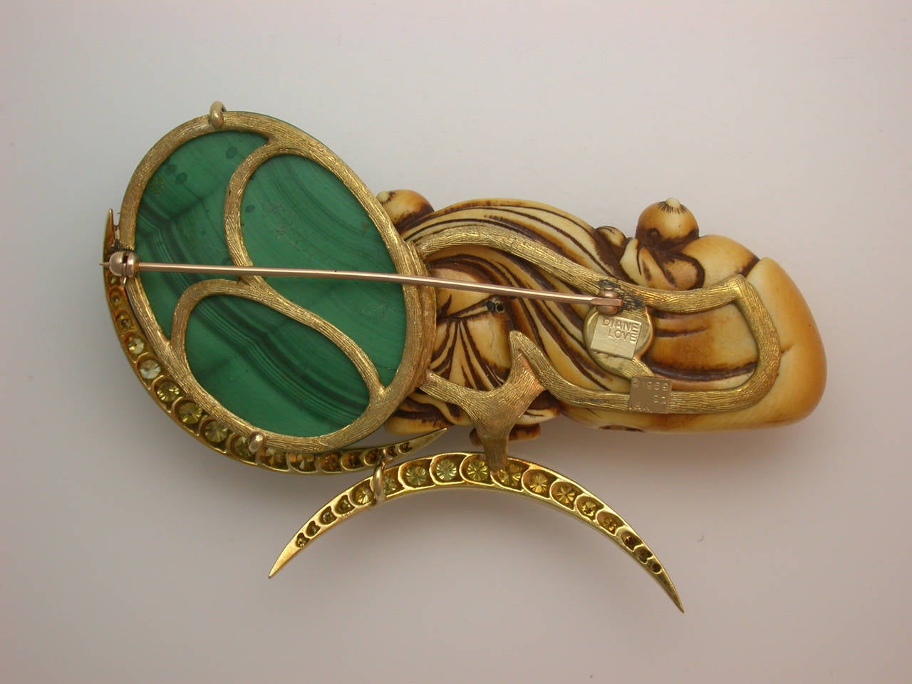 An artistic composition built from disparate elements which work beautifully together, the large pin centering an antique Japanese netsuke, aged to a soft honey yellow, its carved drapery mirrored cleverly by the banding in the bright malachite
