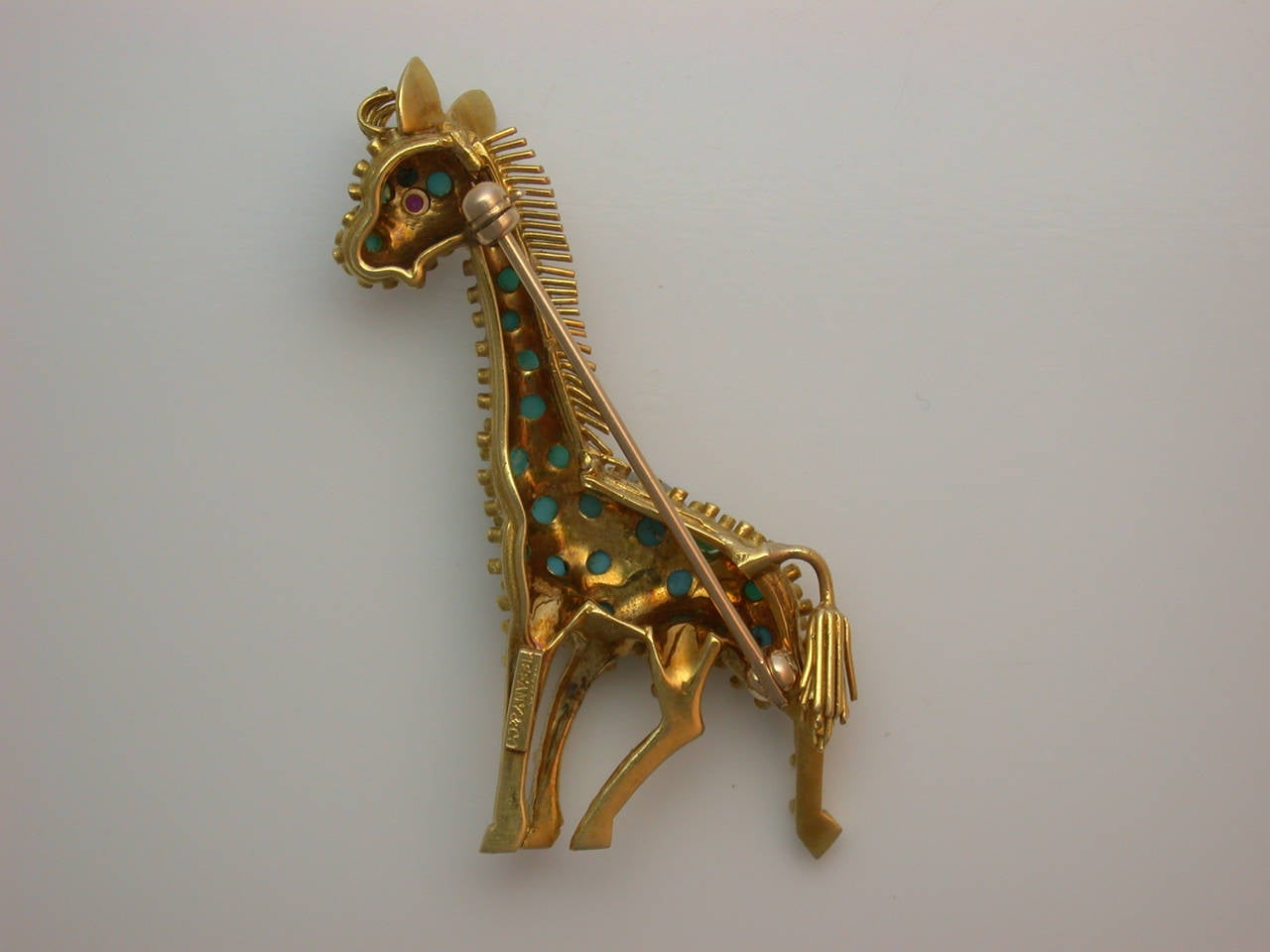The whimsical pin realistically designed as a standing giraffe, its textured yellow gold body set with cabochon turquoise accents, a pleasant contrast to the polished gold ears, mane and tail, further enhanced with a single faceted ruby eye, signed