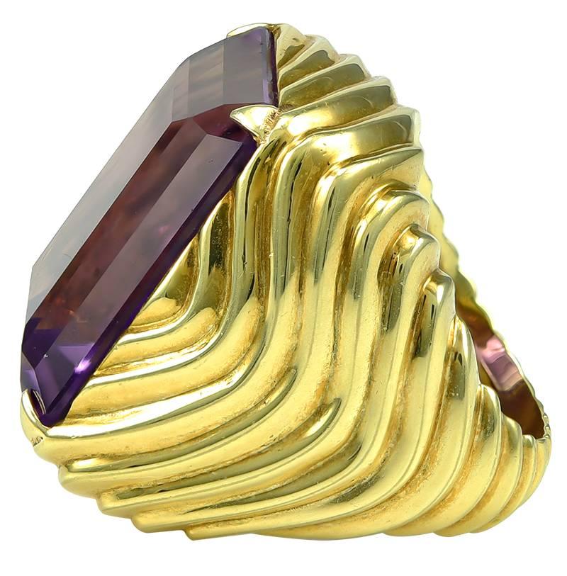 The bold ring centering a rectangular-cut amethyst measuring approximately 22mm by 17mm, weighing approximately 25 carats, atop a polished yellow gold mount decorated on all sides with ribs in waves, stamped 18K, fits finger size 6 to 6 1/2,