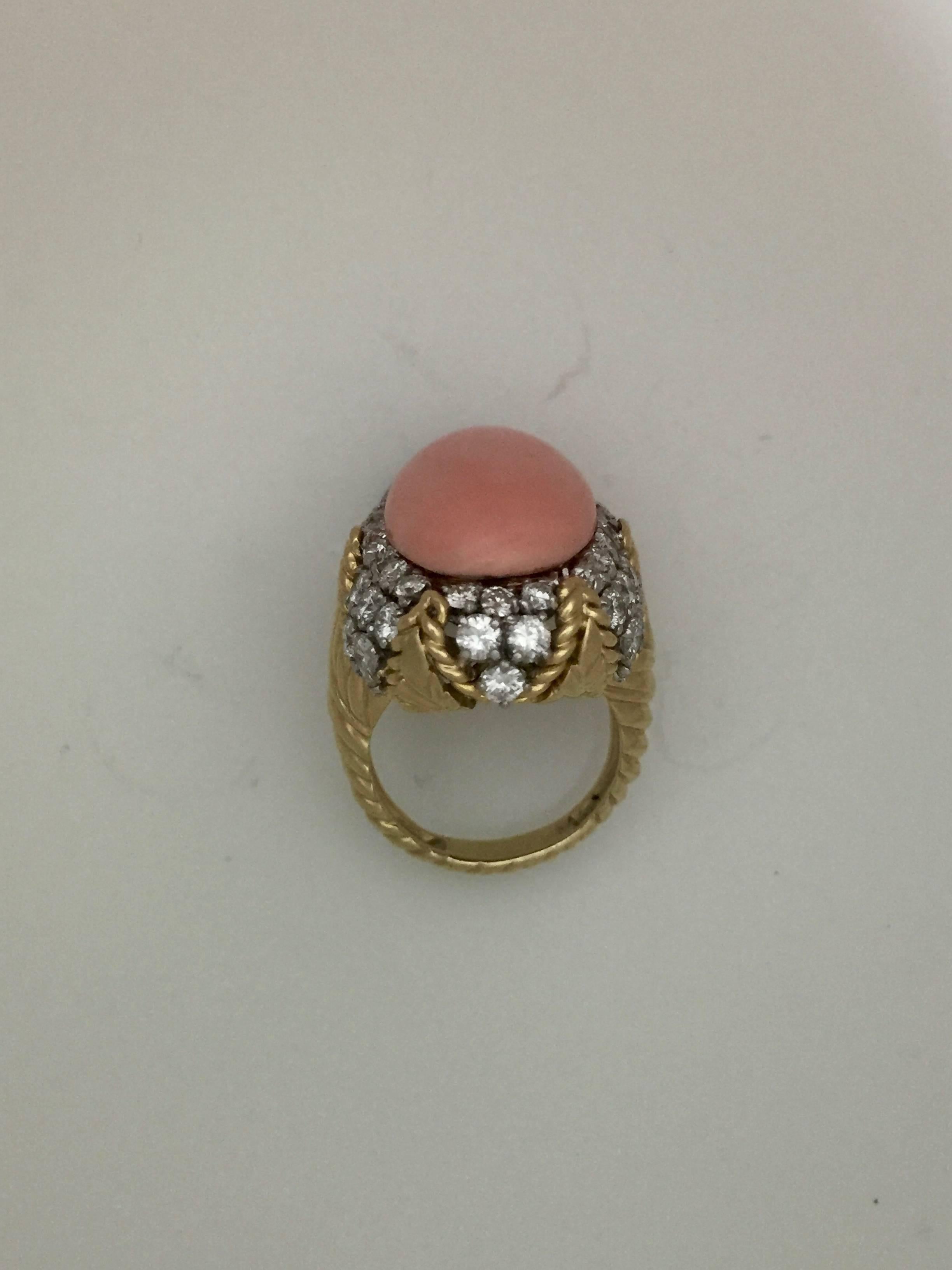 The elegant cocktail ring set along the top with an oval cabochon coral (measuring approximately 20 x 13mm), a flattering feminine shade of medium salmon pink, held from below (i.e. without prongs) above a decorative fluted gold and diamond mount