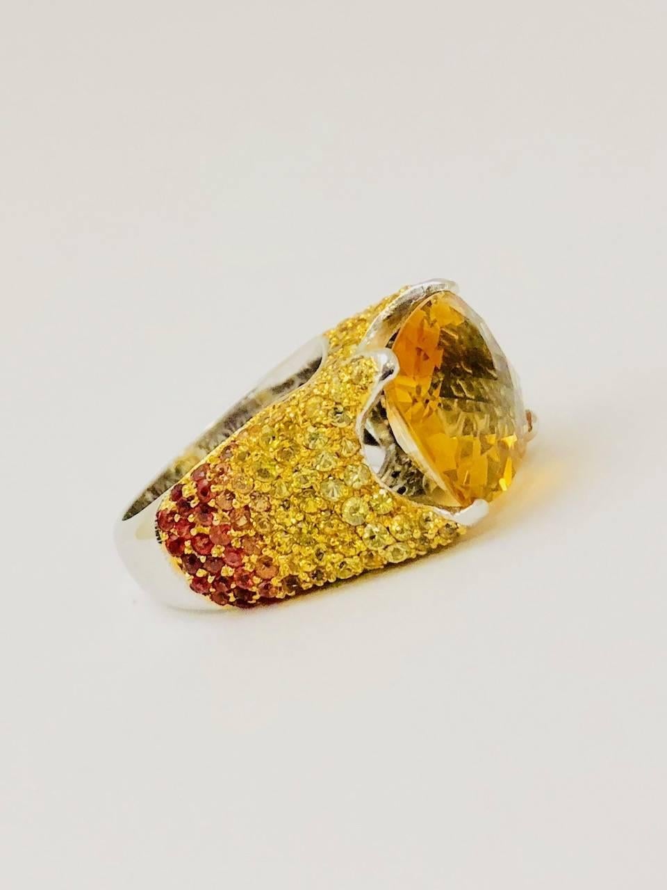 Beautifully crafted in 18 karat white gold, this stunning ring is an eye catcher!  Begin with an East West four prong set oval checkerboard faceted orange citrine.  All 4 sides are encrusted in sapphires that range from the brightest yellow down to