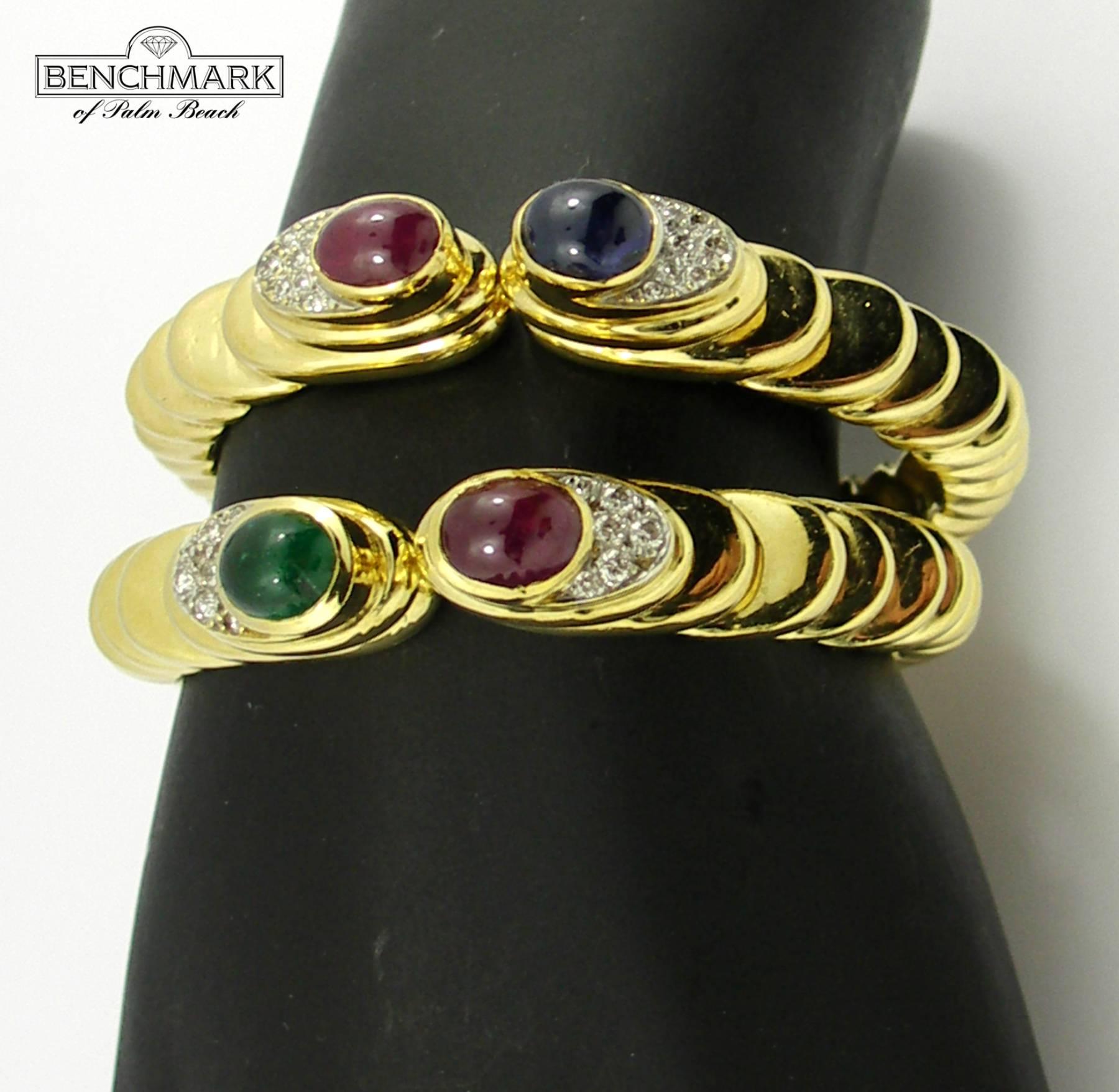 A pair of 18K yellow gold bracelets, of a split front and tiered design. Each bracelet has a bezel set cabochon ruby, and while one bracelet is set with an emerald, the other bracelet is set with a sapphire. The bracelets are also set with a total