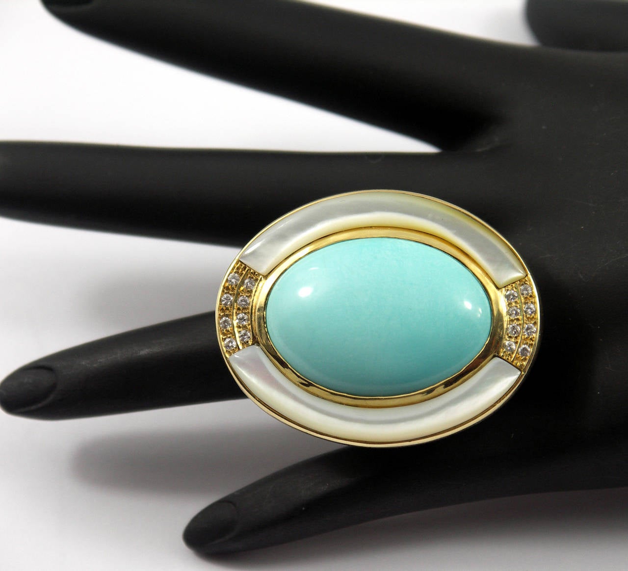 One 18K yellow gold ring measuring 1 3/4 inches long, by 1 1/4 inches wide. Centered around an oval cabochon turquoise measuring 30mm X 22mm. The turquoise is bordered on the East and West by mother of pearl, and bordered by 0.20ct of round