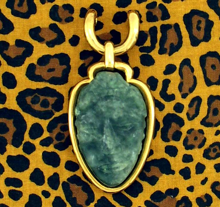 A sleek 18K yellow gold pendant, with an outline resembling an arrowhead. Inside the framework is a carved jade face resembling that of Mayan or Aztec art.
