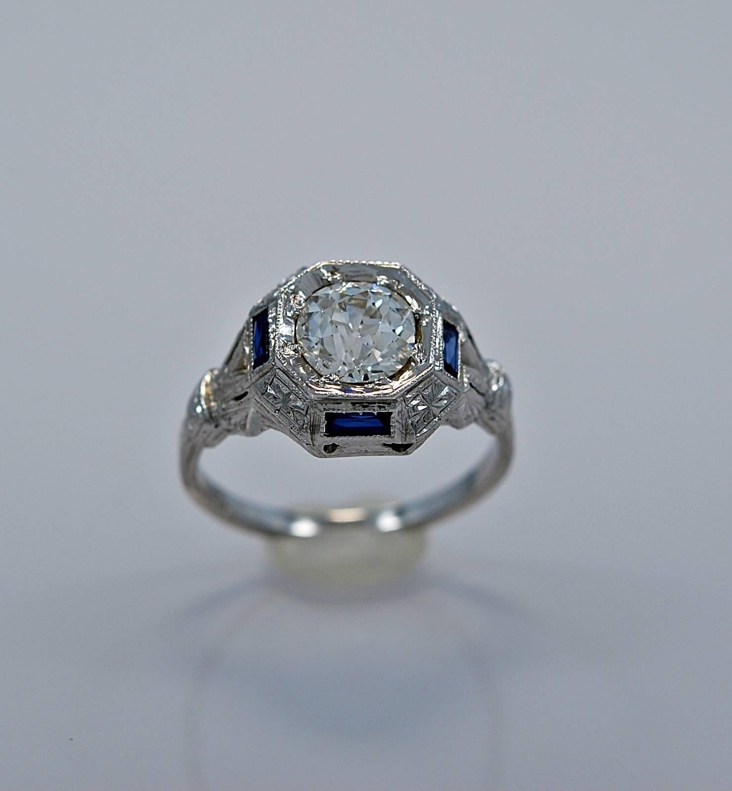 An Art Deco 18K white gold & diamond engagement ring featuring a 1.00ct. apx. diamond of VS1 clarity and J color. Four synthetic sapphires (consistent with the time period) accent the center diamond on each side of the head. This ring is beautifully