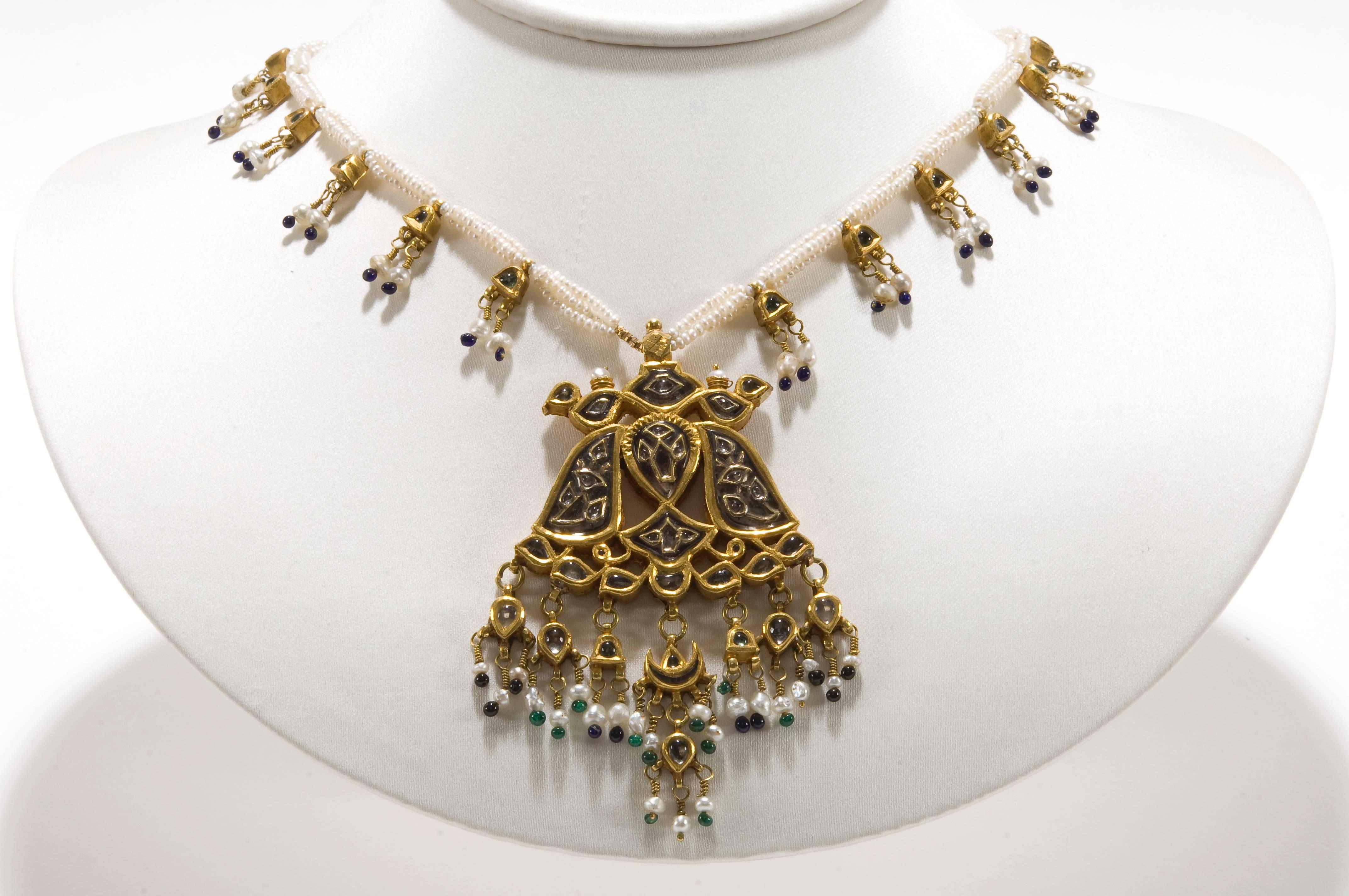 A gold pendant, JUGNI, kundan set with foiled rock crystals, embellishes with carved and gold floral pattern. The reverse of the pendant is decorated with a flower and bird design in embossed sheet gold. The pendant is suspended from three strings