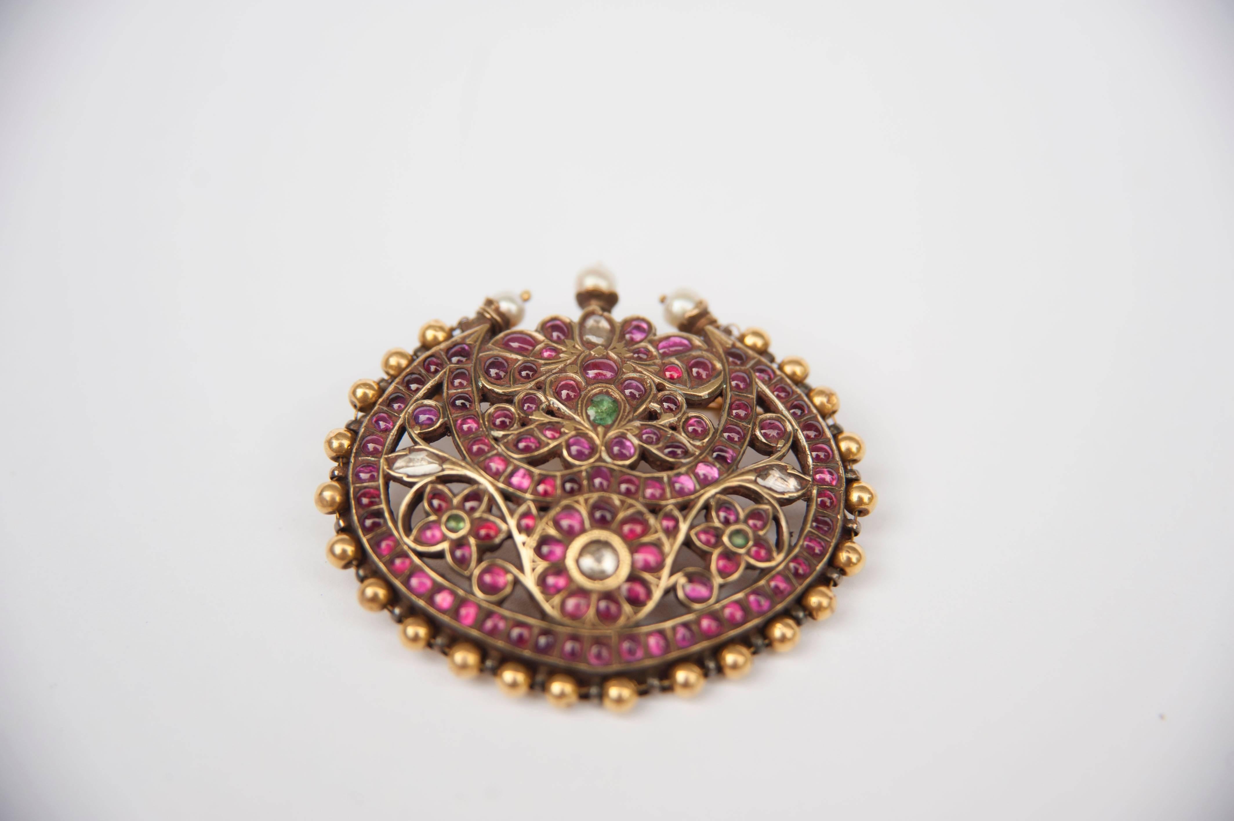 A head ornament, RAKODI, consisting of an open worked gilded silver round pendant, depicting a floral and leaf design, the whole kundan set with rubies, diamonds and foiled green beryl, crowned with three pearls, and a semicircular decoration of