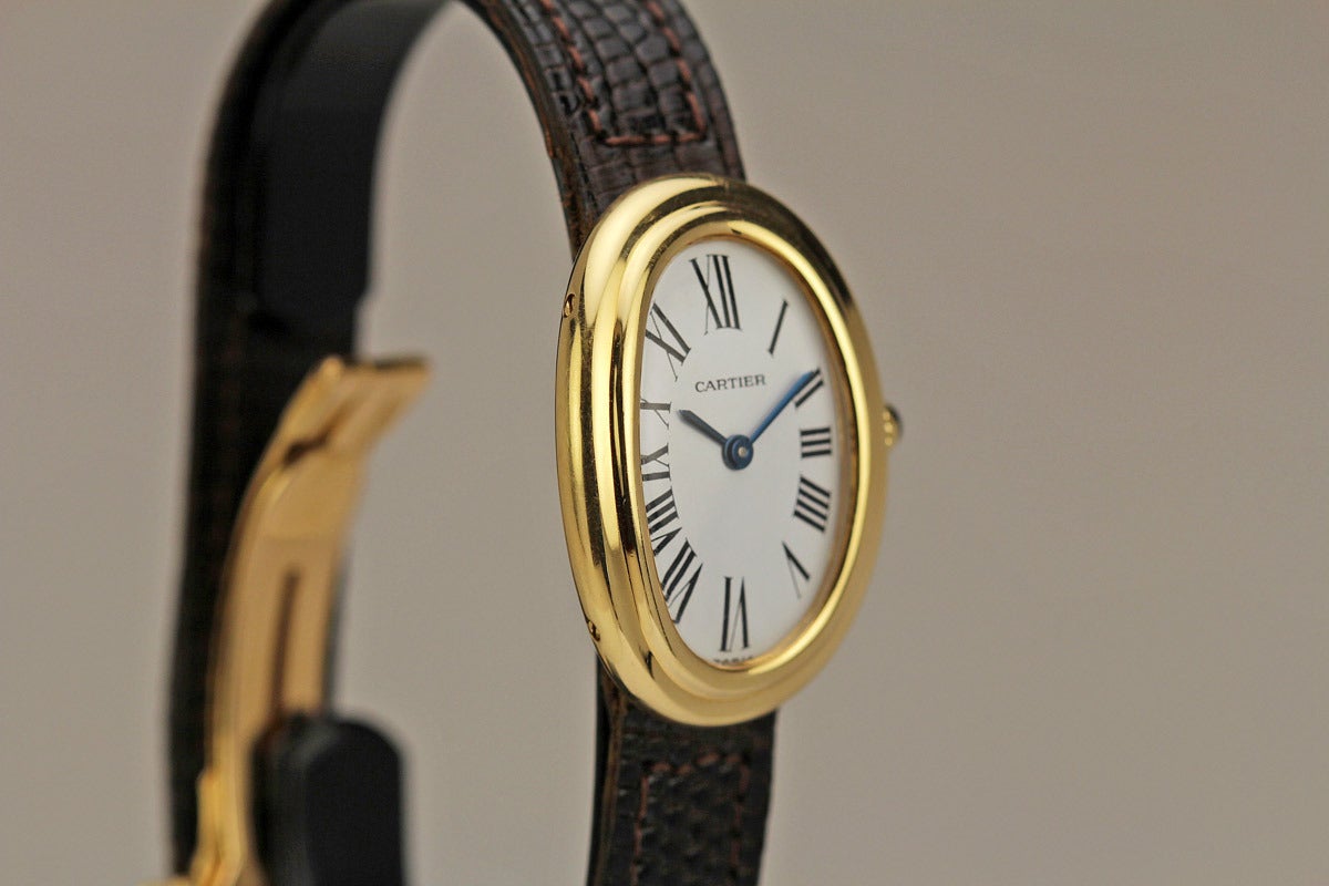 Timeless Cartier Baignoire lady's wristwatch with a matte finish dial, Roman numerals, blued steel hands, curved oval gold case with hidden lugs on a leather strap and deployant Cartier clasp.