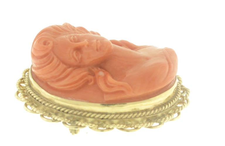 This brooch features beautifully carved salmon coral. The carving is of a woman's face and neck. The large coral carving is set in a 14-karat yellow gold frame. The brooch measures 1.7 inches in length, 1.5 inches in width, and 20 millimeters in