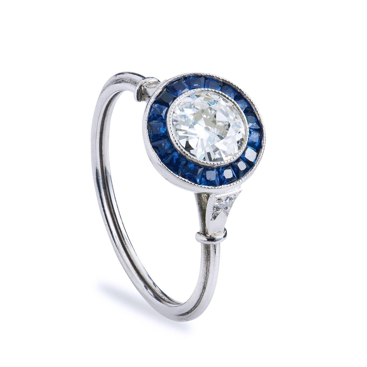 Crafted in platinum, this ring features a .66ct H/I VS graded old European cut center diamond surrounded by .70ct of caliber cut channel set sapphires. The ring features a thin shank for a very soft and feminine feel that is set with pave diamonds. 
