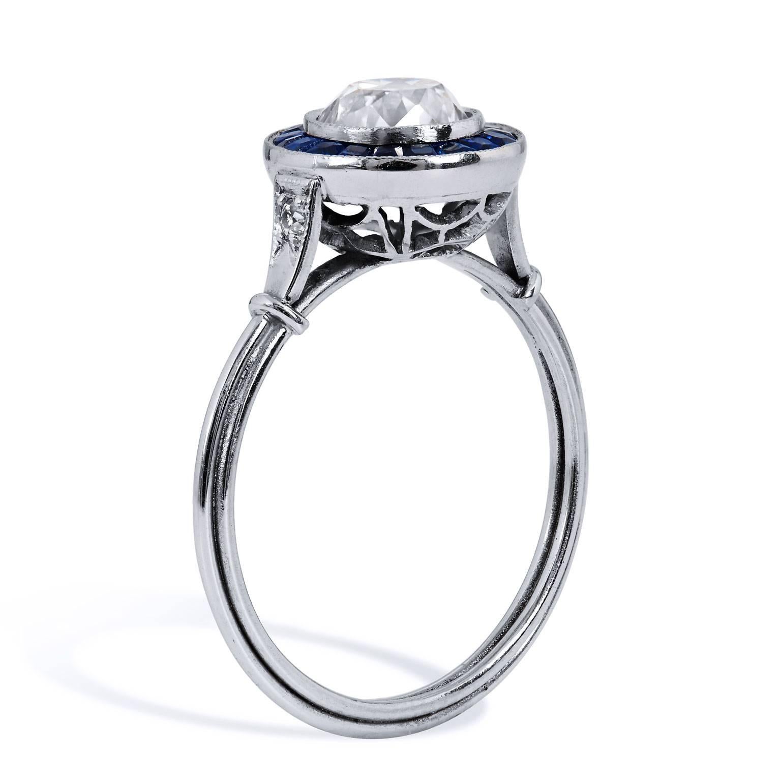 Crafted in platinum, this ring features a 1.16 carat L/M/SI1 graded Old European cut center diamond surrounded by 0.70 carat of caliber cut channel set sapphires. The ring features a thin shank for a very soft and feminine feel that is set with pave