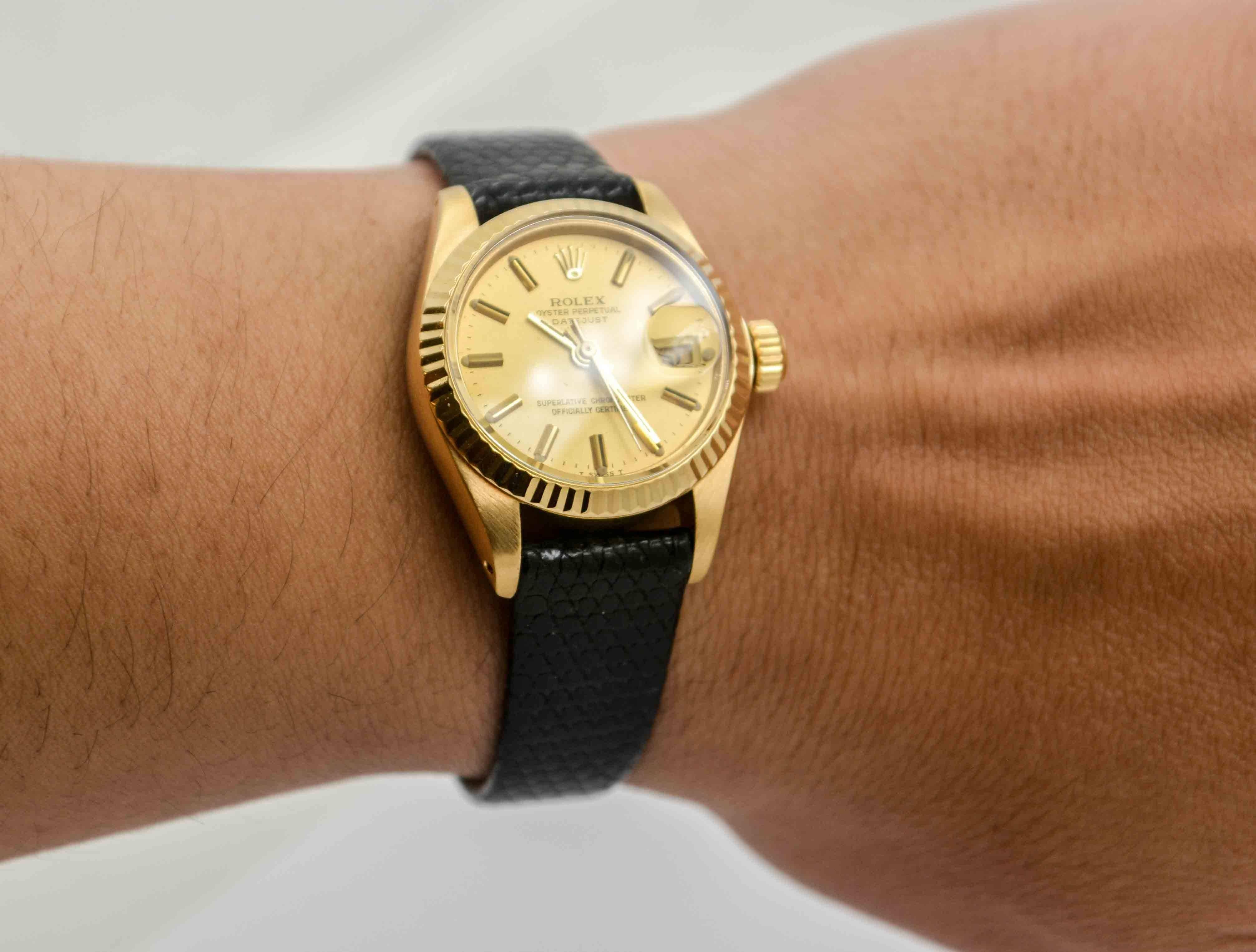 A classic ladies 18kt. yellow gold 26mm Rolex Datejust, Champagne index dial watch with an acrylic crystal. Automatic movement, circa 1979. Black leather band.