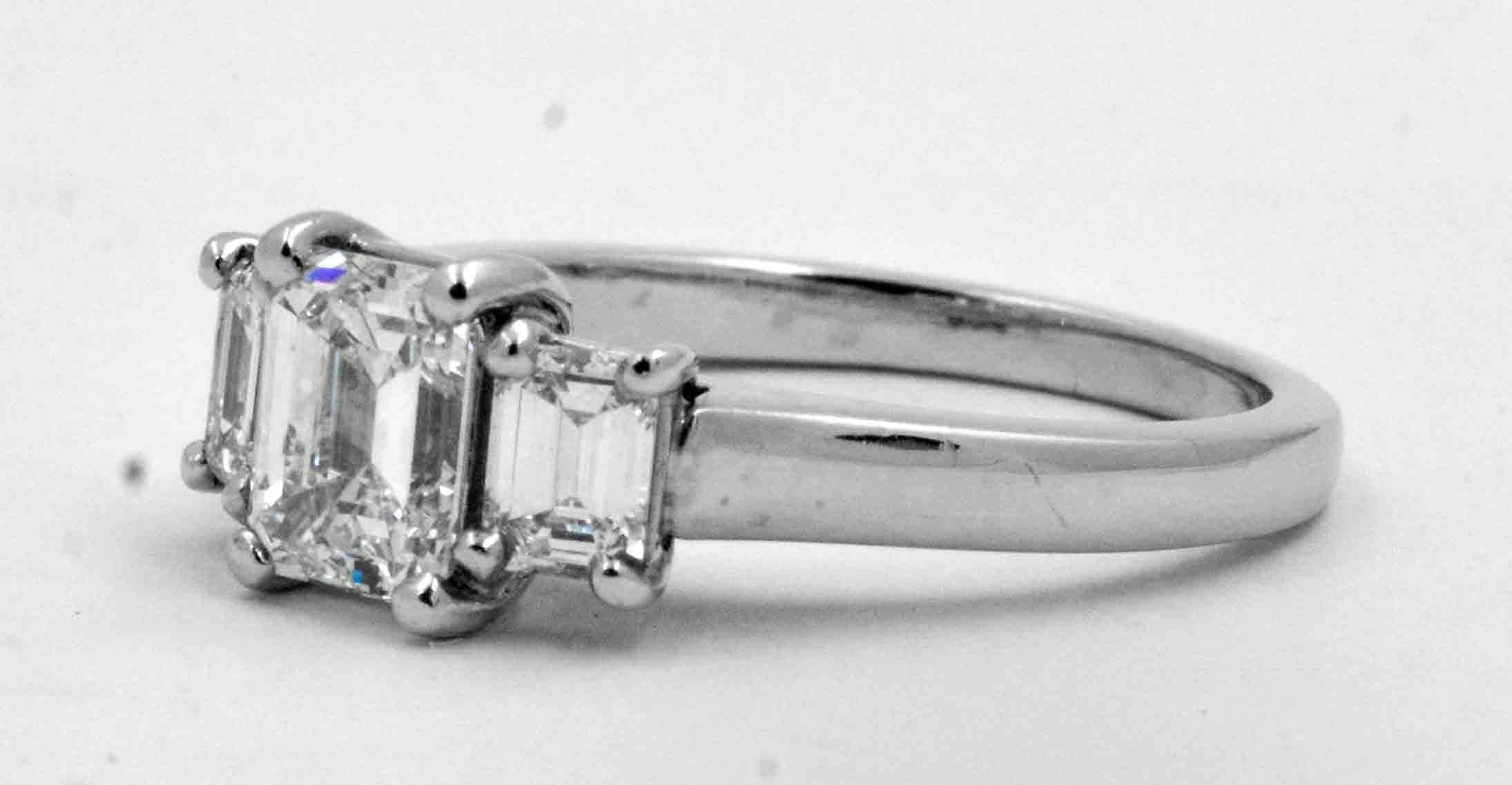 A luxurious emerald cut 1.00 carat diamond gets center attention in this attractive classic three stone engagement ring crafted in scintillating, highly polished platinum. The opulent emerald cut diamond weighs exactly 1.00 carat  (F color and VS1