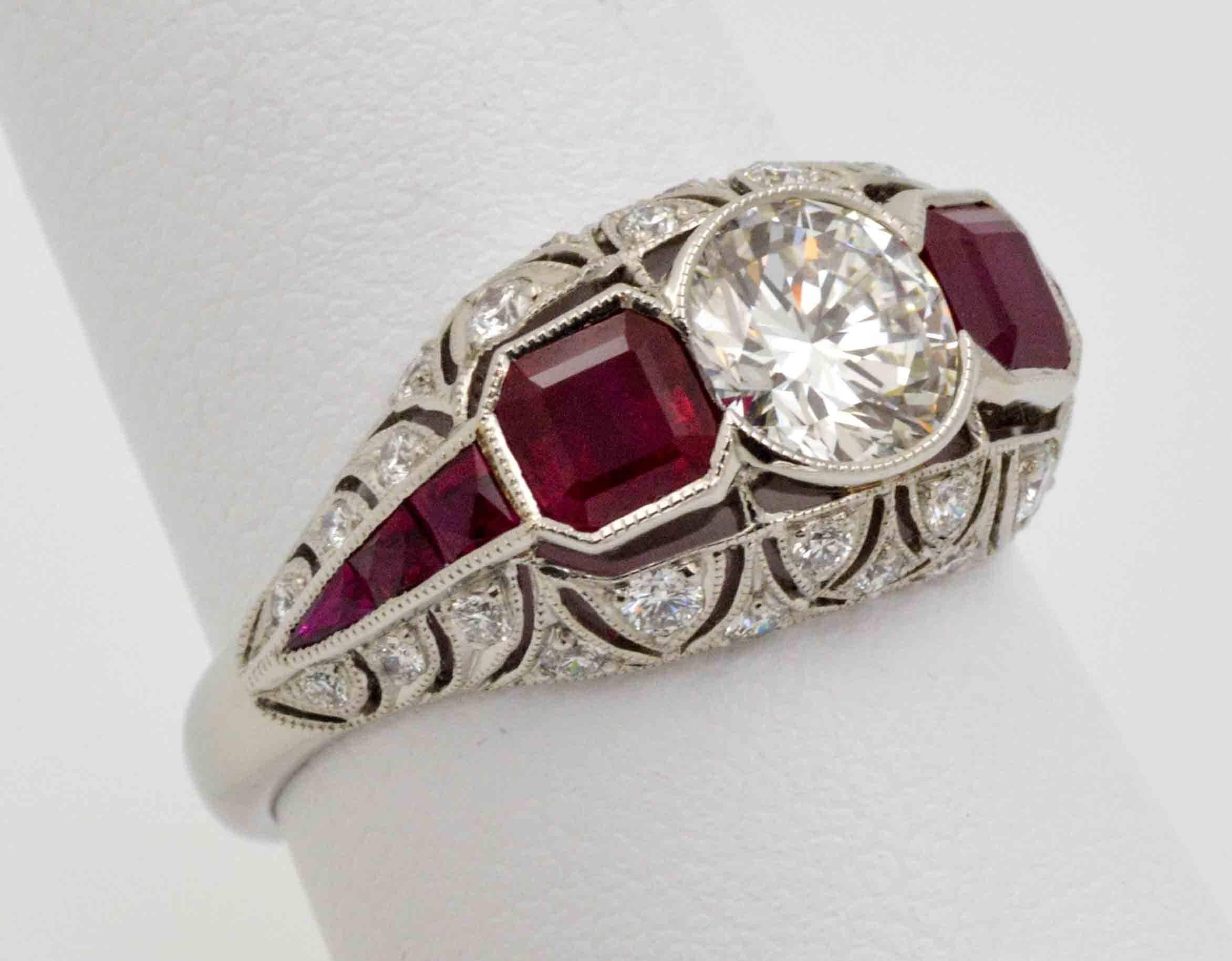 Inspired by Art Deco Style design, this custom handmade engagement ring features an approximate 0.94 carat round brilliant cut diamond with J color and VS1 clarity. The ring is accented with two cushion cut bezel set rubies weighing approximately