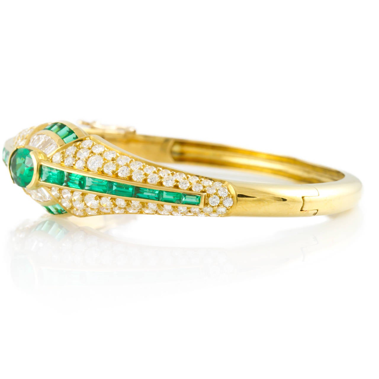 Lady's 18K yellow gold hinged bangle bracelet with two figure eight safeties, half bezel-set with one (1) Oval Mixed cut Emerald, channel-set with fourteen (14) Baguette cut Emeralds, channel-set with ten (10) Square Step cut Emeralds, channel-set