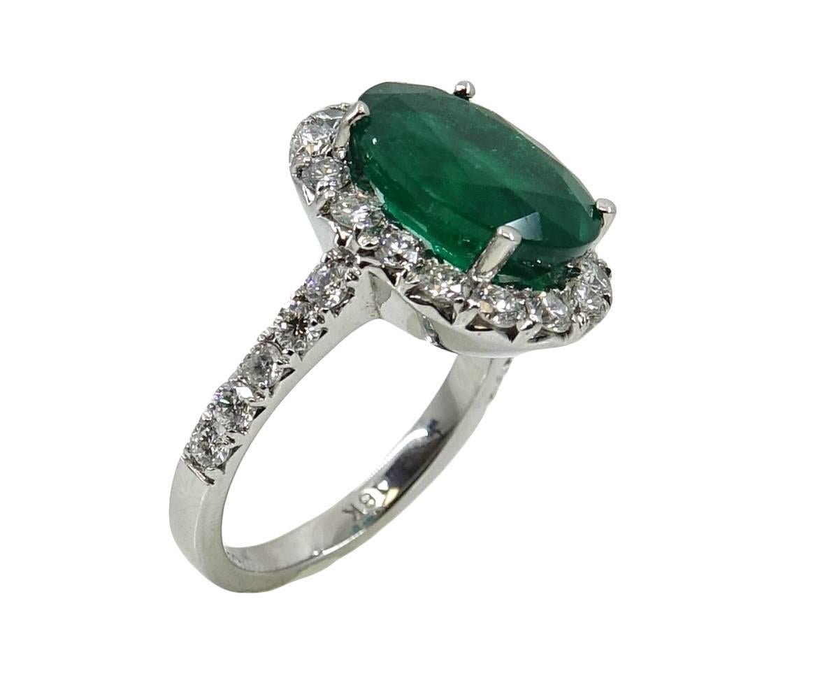 This Classic Style 18K White Gold Ring Has A Vibrant Oval Cut Emerald Stone In The Center Weighing A Total Carat Weight Of 5.20 Carats. Round Diamonds Outline The Emerald and Weigh A Total Carat Weight Of 1.20 Carats. This Beautiful Ring Is A Size 7