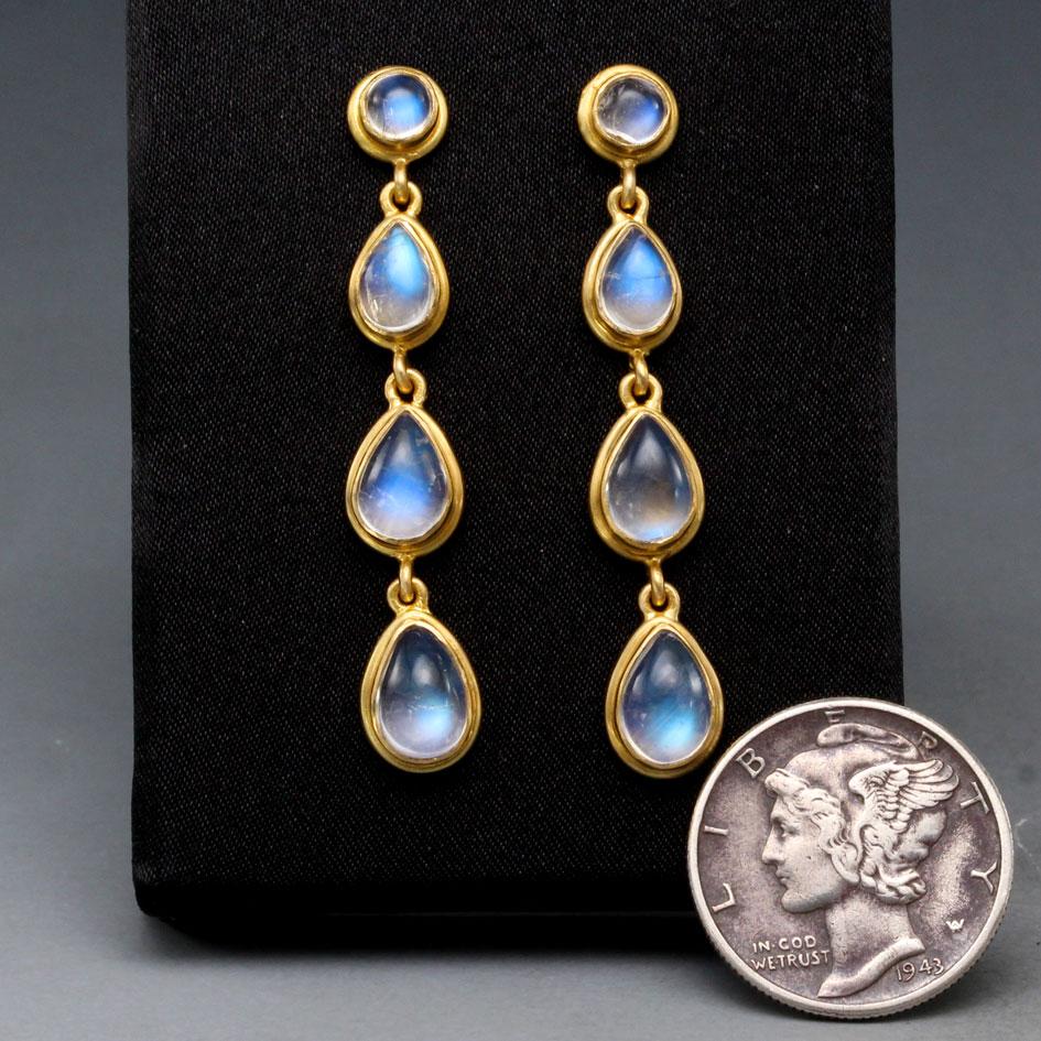 Abgestufte, birnenförmige Cabochons (4 x 6 mm bis 6 x 8 mm) aus schimmerndem Regenbogenmondstein sind in reichhaltige, abgerundete, doppelte Lünettenfassungen gefasst und baumeln in diesem Design unter 5 mm großen Regenbogenmondsteinpfosten.  Das