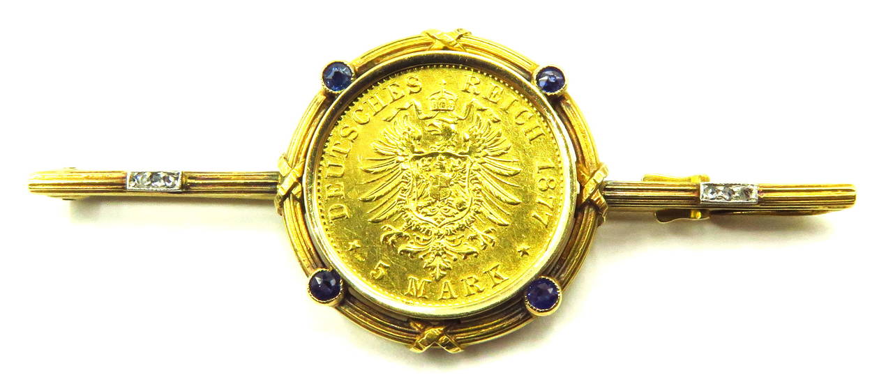 German coin, 5 Marks Dated 1877, made into bar pin. The bezel is accented with 4 bezel set sapphires. and the sides are accented with small platinum bars of 3 rose cut diamonds. I left the patina on this pin, but will be glad to clean it up if you