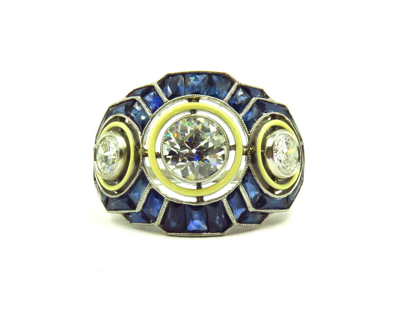 This elegant deco style 3 stone diamond ring is surrounded by french calibre cut sapphires. Each of the diamonds is first surrounded by off white color enamel. The center diamond weighs approximately 1ct. The 2 side diamonds total about  .70ct. This