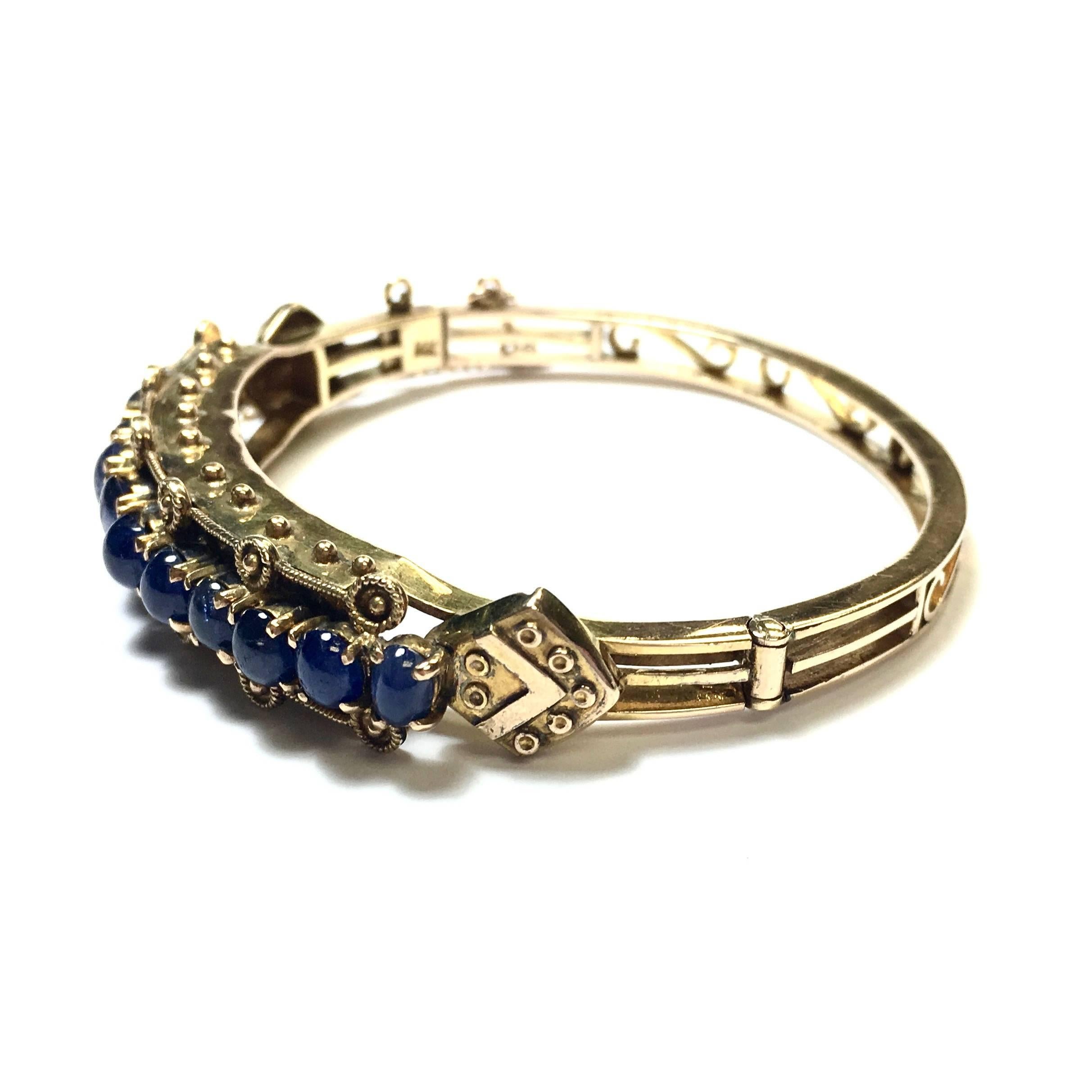 Gorgeous  hinged bangle bracelet crafted in 14K yellow gold, featuring 12 graduating cabochon cut blue sapphires  set on the front, suuported by elaborated gold sections with beadwork, completed by open scroll design 5 mm wide back . 
Apprximate
