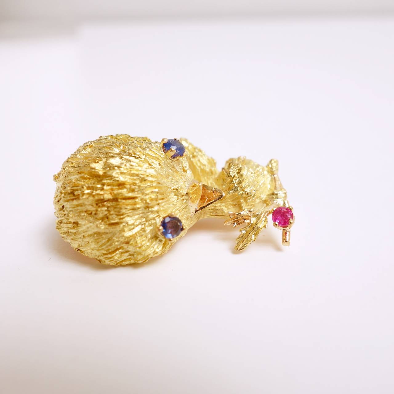 Sweet 18K yellow gold textured finish bird brooch with blue sapphire eyes and a round ruby accent. 
Measurments: 32 mm H x 22 mm W x 16mm x 12 mm D 
Weight: 13.6 grams