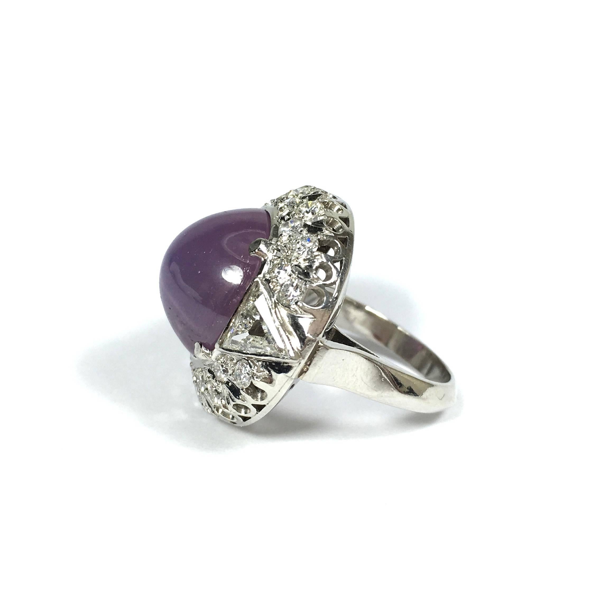 This gorgeous one-of-a-kind platinum ring features an approximately 16ct  round cabochon natural, translucent purple-pink sapphire flanked by two beze set trillion cut diamonds, supported by a bezel of diamonds, completed by a three millimeter wide