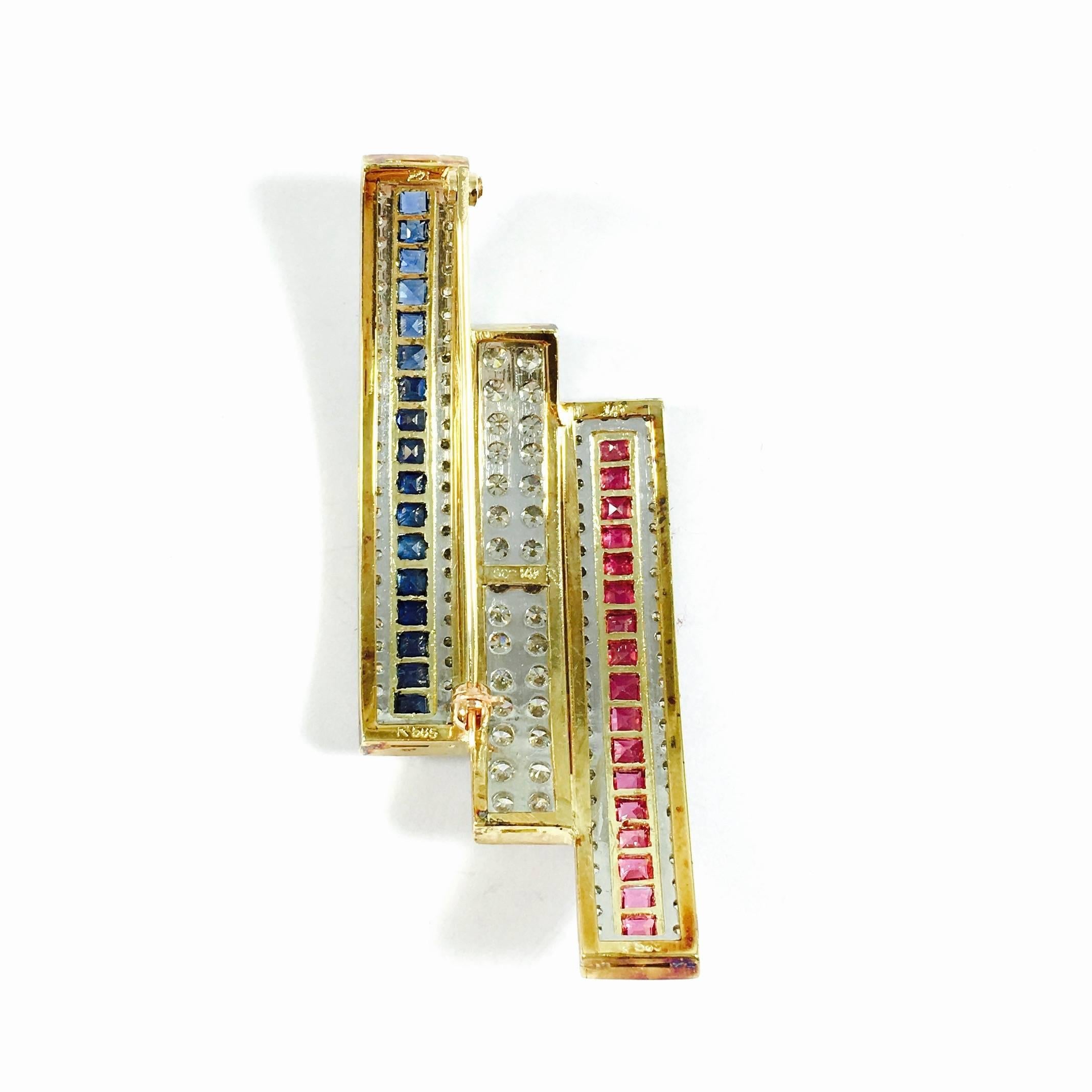 Featuring a unique, curved design, the pin is composed of three 14K gold bars set with rubies, diamonds and blue sapphires. 
17 square cut natural rubies: approximate total weight of 1.00 ct
17 square cut natural blue sapphires: approximate total