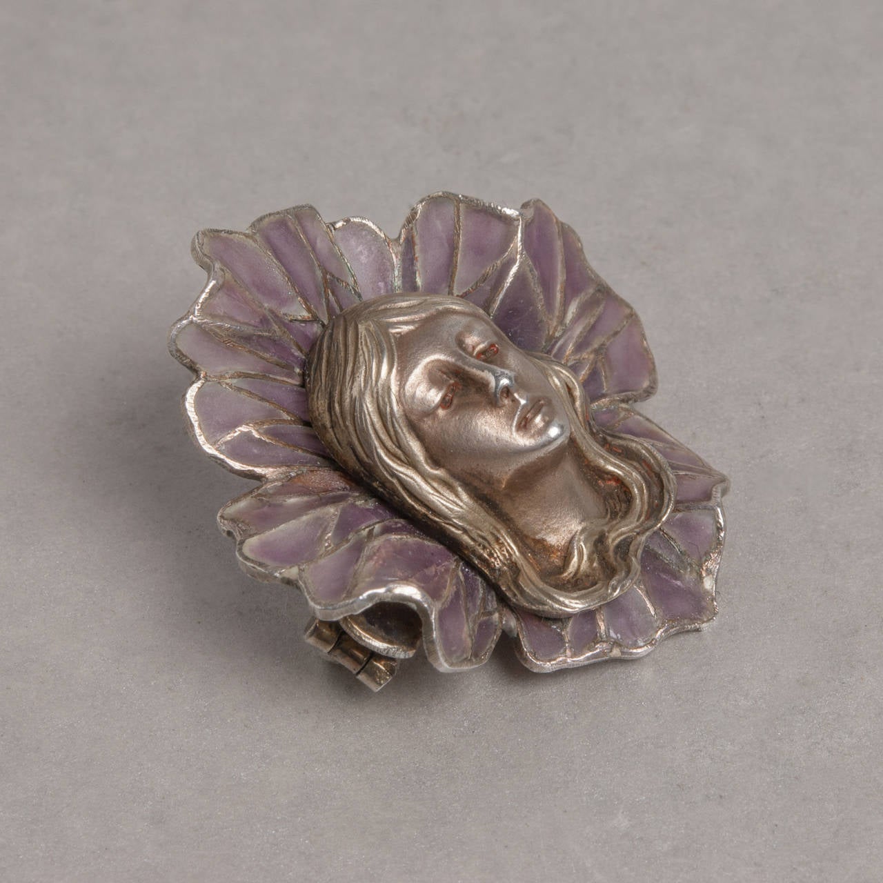 A stunning silver and plique-à-jour enamel pin depicting a woman's face surrounded by flower petals. The floral plique-à-jour enamel work creates a unique effect due to its subtle violet hue and semi-translucence appearance.