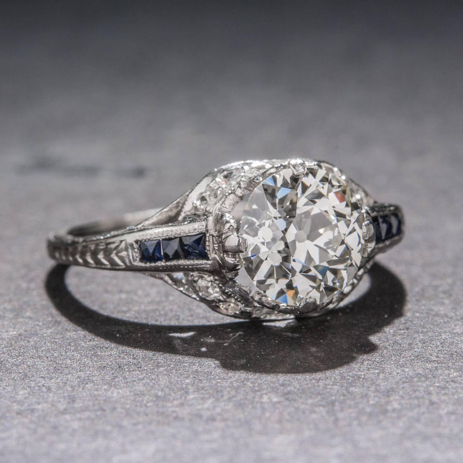 An elegant Art Deco ring with a beautifully crafted and elaborately detailed platinum setting. The Old European cut center diamond weighs 1.44 carats and has a channel of sapphire on each side. This ring is currently size 6.75.

