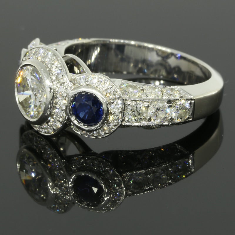 The ring features one center round brilliant cut diamond weighing 0.83CT. The diamond is an I in color and an I/I1 in clarity. The ring also features two round brilliant cut, natural blue sapphires weighing 0.73CTW. These three main stones are bezel