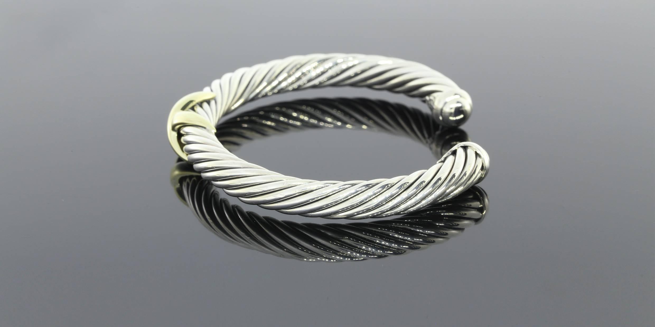 This cuff is part of the designer line David Yurman. The cuff features a cable style base with a yellow gold X detail. The cuff measures 7 millimeters in thickness. The bangle appraises for $775.