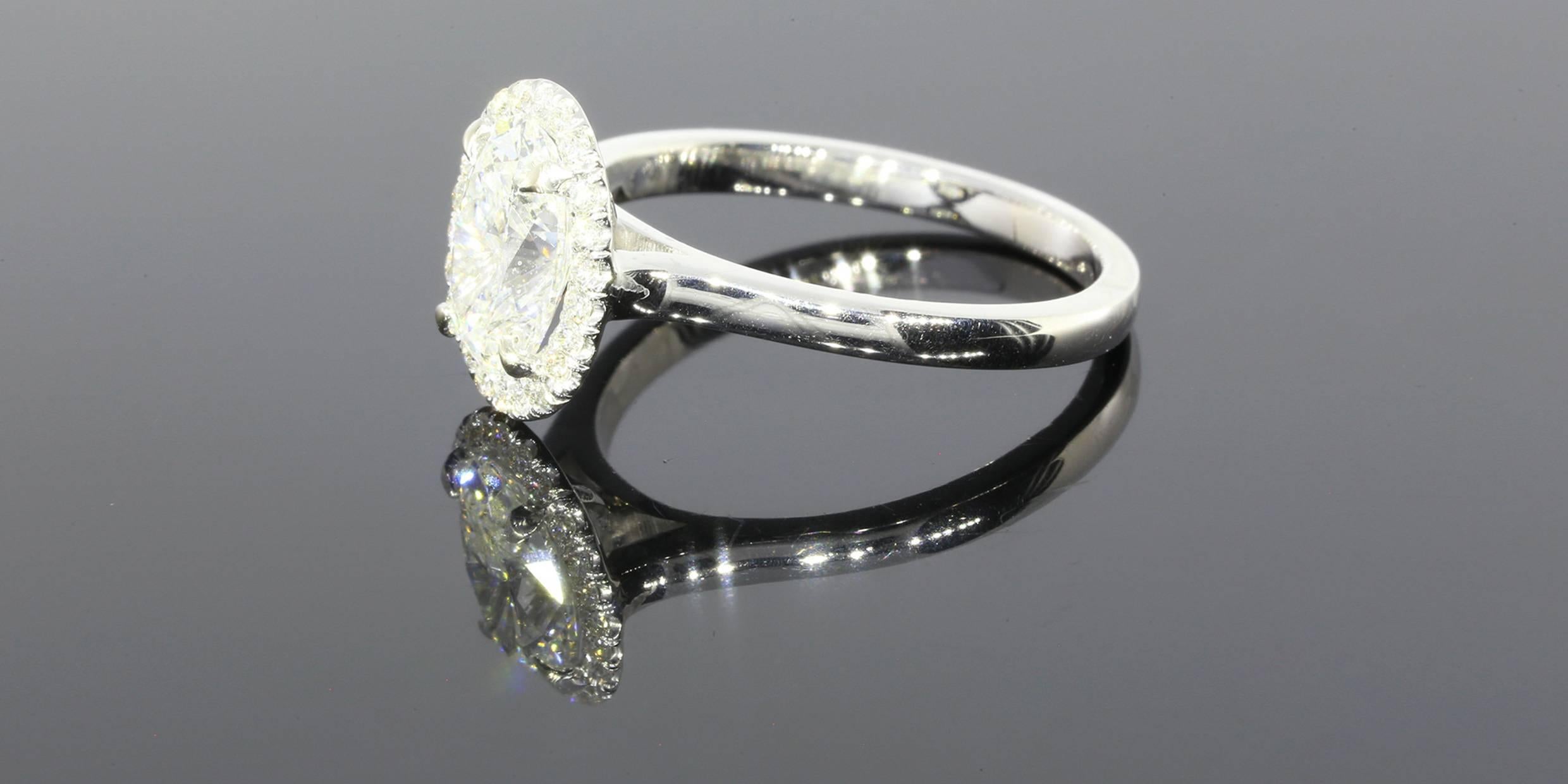 The ring features one brilliant cut oval center diamond weighing 1.41 carats. The diamond has a GIA lab report, number 6147600273. The diamond is an E/VS2 in quality. The diamond is set in a 14 karat white gold halo style mounting featuring round
